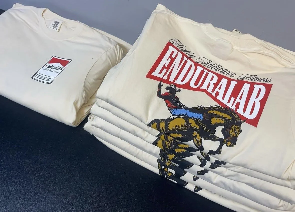 Stack of printed white T-shirts, one showing a graphic with a stockyards inspired design for a local gym.