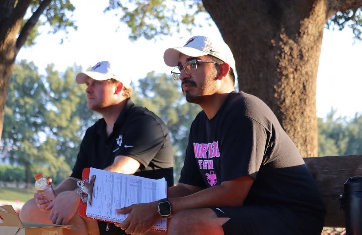 Two men sitting on a bench under a tree during daytime. The man on the right is wearing glasses, a black T-shirt with pink writing, a white cap, and is holding a clipboard and a water bottle. The man on the left is wearing a black polo shirt and a white cap, and is looking ahead. Background shows green trees and sunny weather.