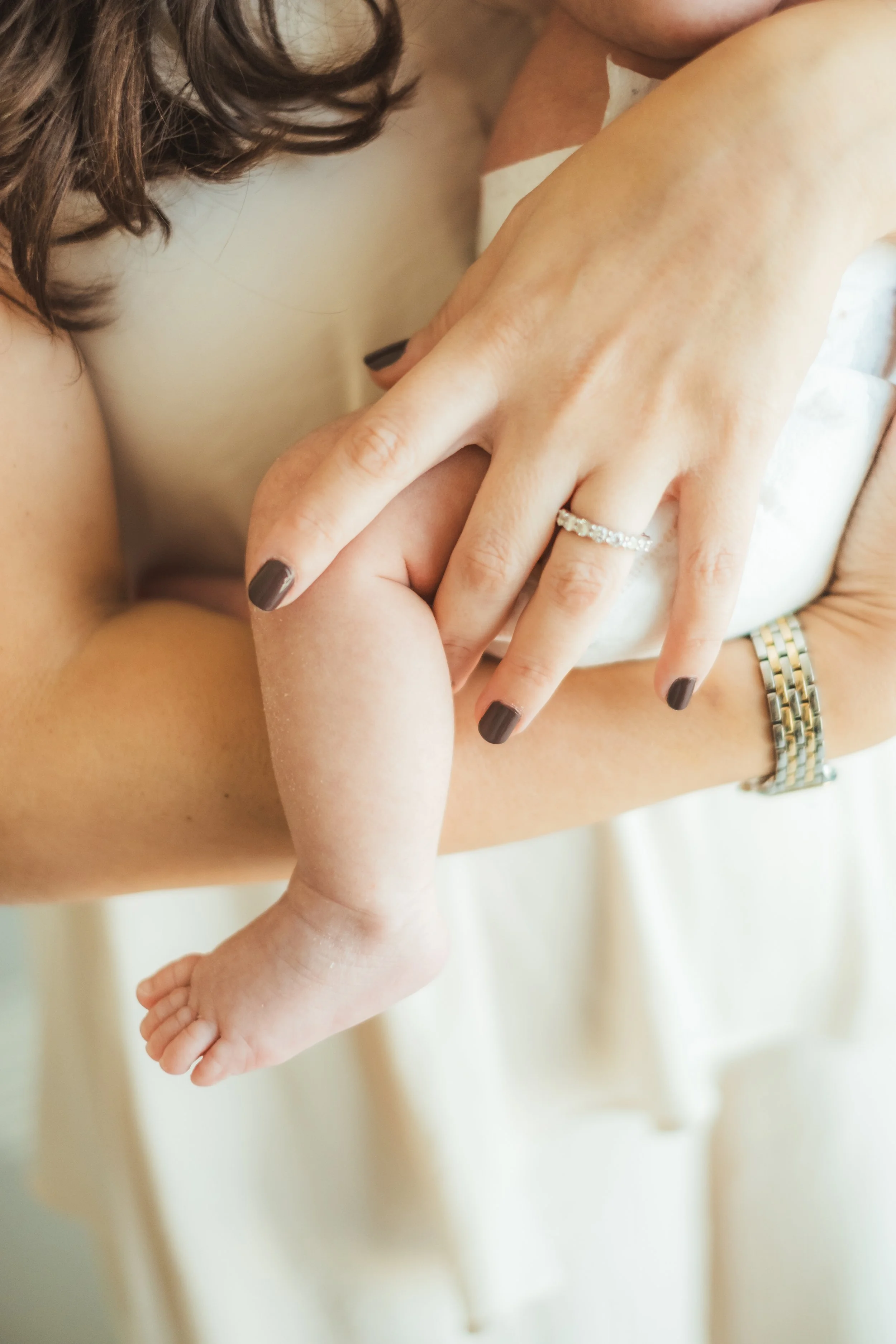 Close-up of a woman's hand showing a ring, gently holding a baby's hand and foot, with a cream-colored background.