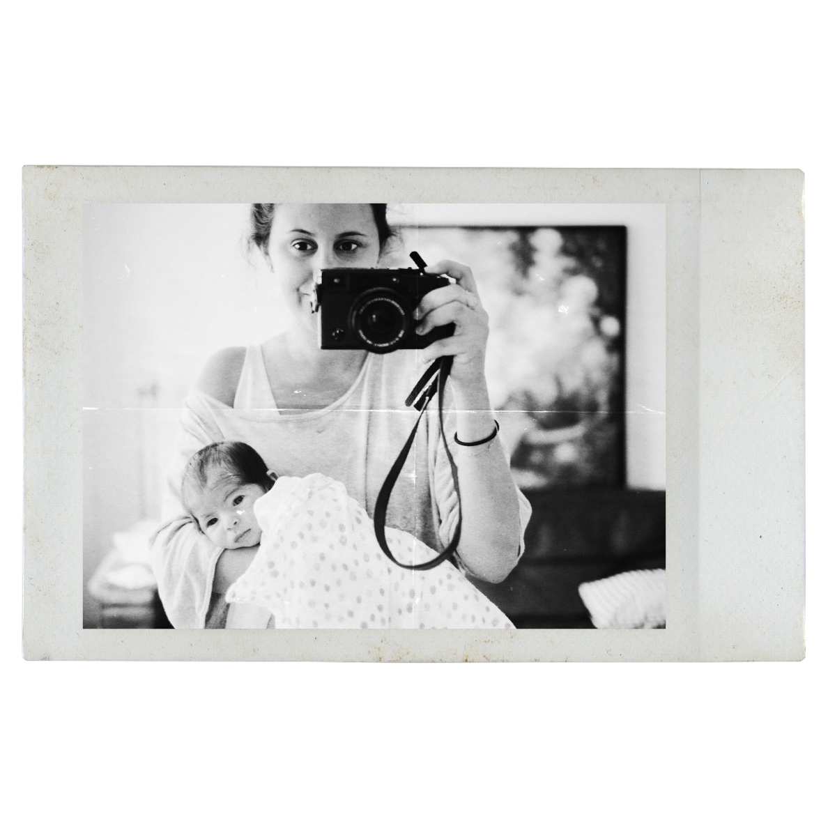 Black and white photo of a young woman taking a selfie with a camera, holding a sleeping baby wrapped in a polka dot blanket; blurred background with a window and outdoor scenery.