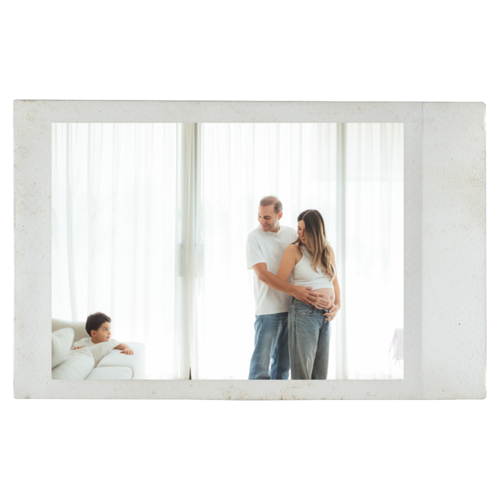 A pregnant woman and her partner smiling inside a bright, white room near a large window, with a young boy sitting on a sofa watching them.