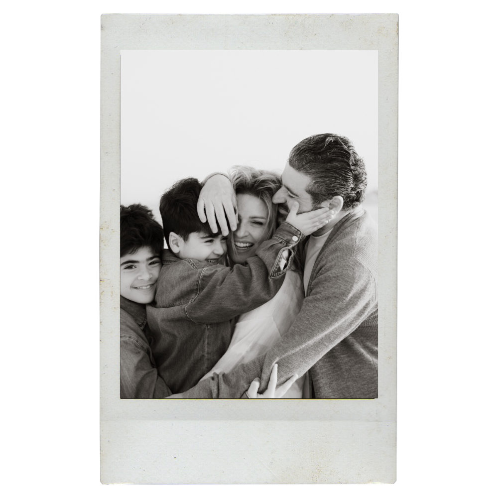 A family of four hugging and smiling in a black and white photo. The mother, a blonde woman, is being embraced by her two young sons and her husband.