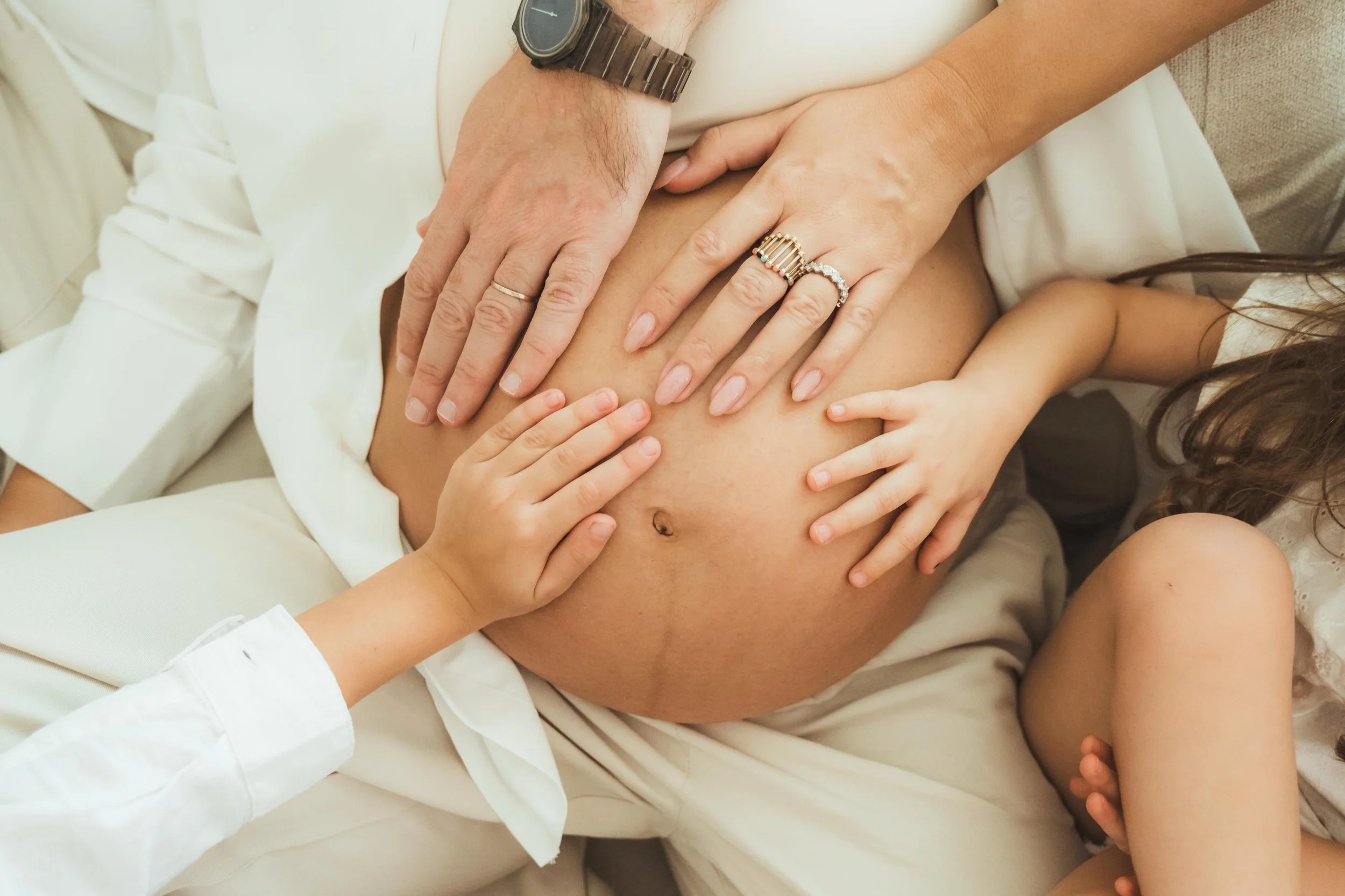 Multiple hands placed on a woman's pregnant belly, with a child touching the belly, all wearing rings and jewelry.