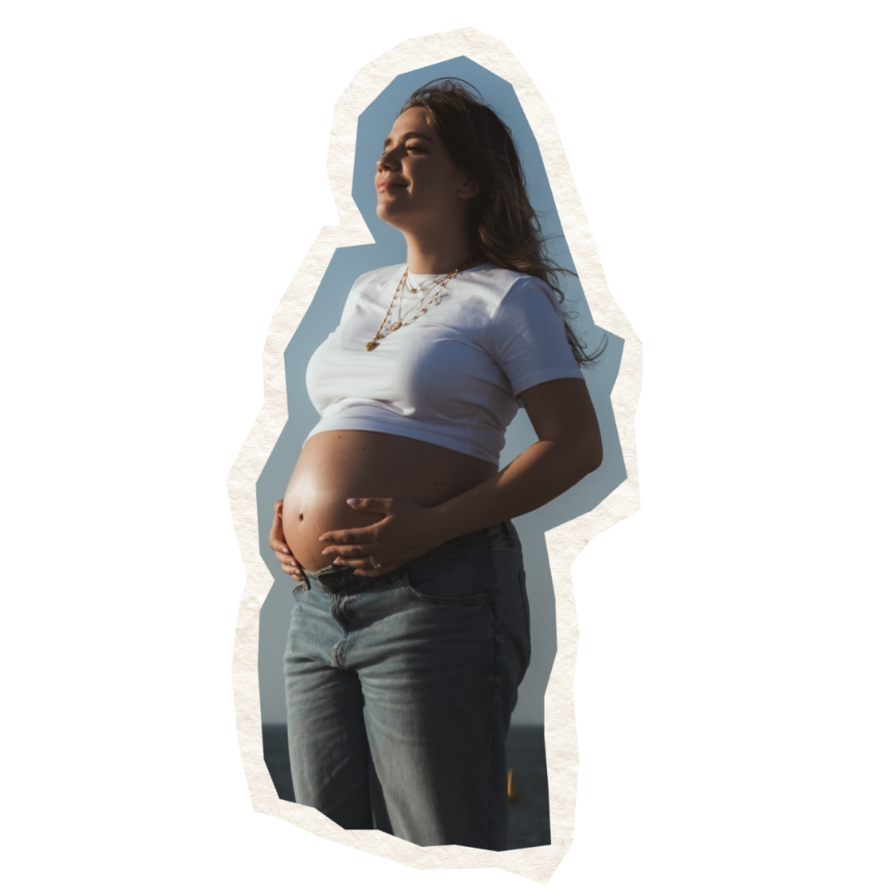 A pregnant woman is standing outdoors with her eyes closed, wearing a white t-shirt and jeans, and holding her pregnant belly with both hands.
