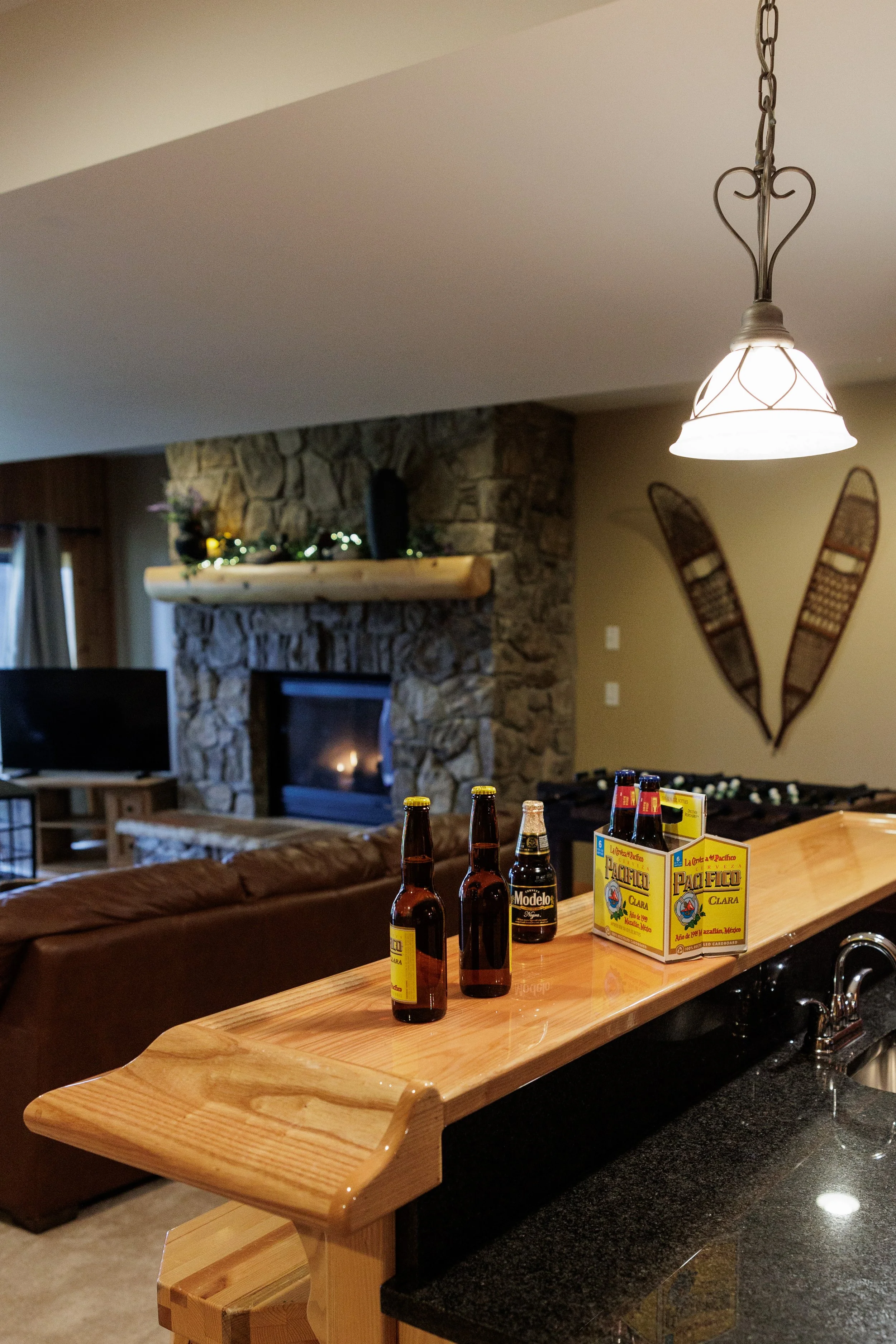 A cozy living room with a stone fireplace and a wooden mantle decorated with Christmas garland and lights. There is a TV and a leather couch visible. In the foreground, a bar counter made of light wood has bottles of beer and a cardboard pack of beer bottles.
