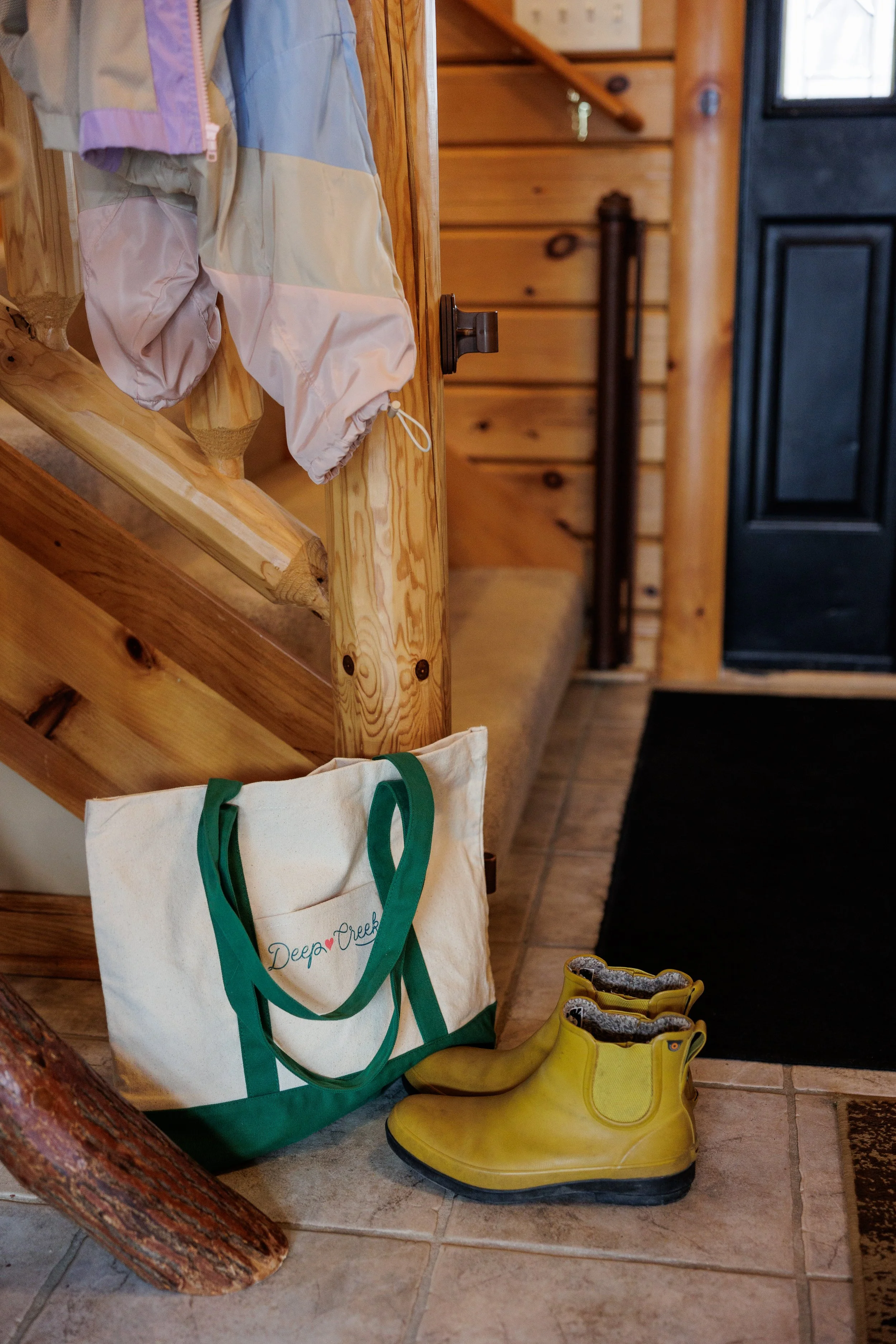 A white tote bag with green handles and the word 'Deep Creek' on it, yellow rain boots, a pair of shoes, and a coat hanging on a wooden stair railing inside a log cabin.