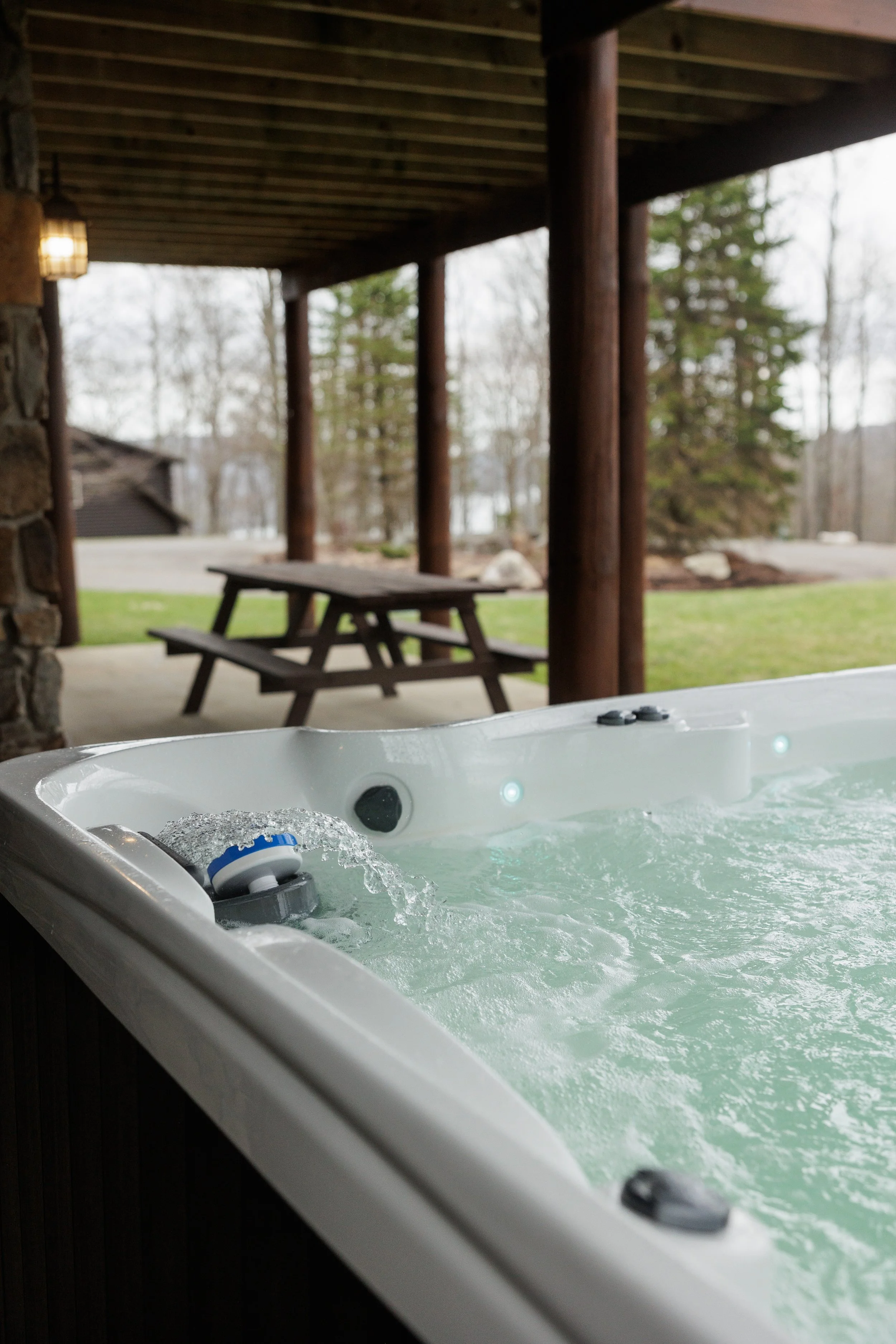 Hot tub on a porch with a view of a yard and trees in the background, some rocks and a picnic table are visible.