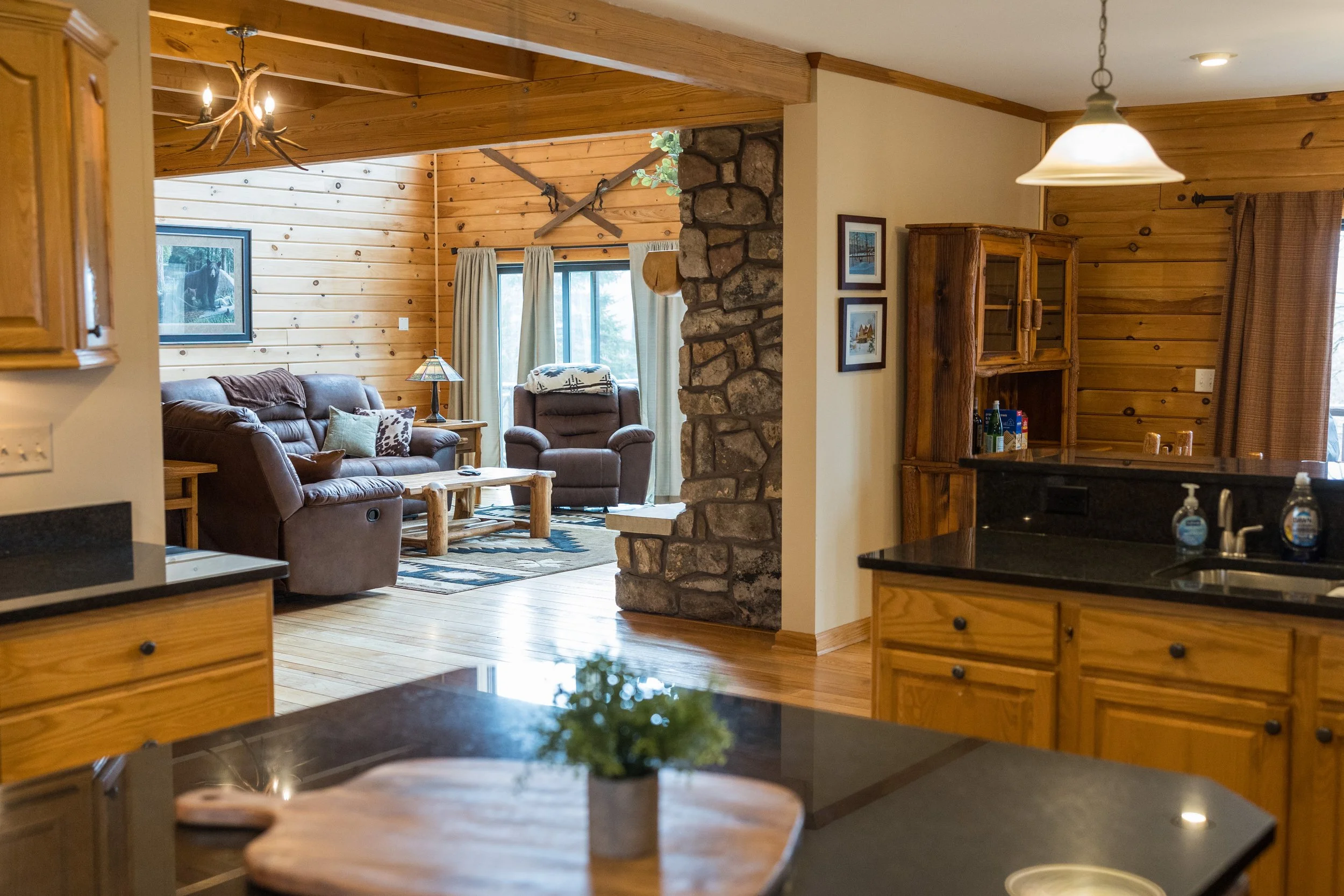 A cozy living room with wood-paneled walls, a stone chimney, and rustic decor, including a framed picture of a bear, a side table with a lamp, and comfortable leather furniture near a large window with curtains.