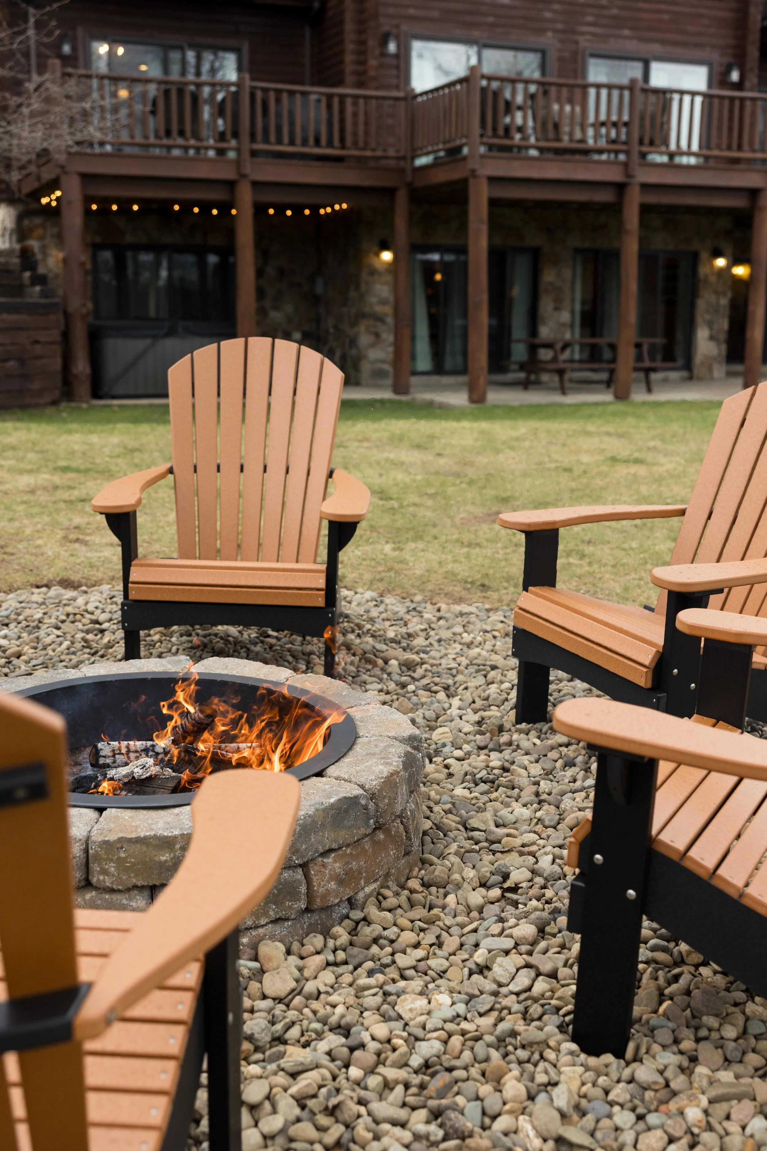 Outdoor patio with Adirondack chairs surrounding a lit fire pit in a backyard, with a multi-story wooden house with large windows and a balcony in the background.
