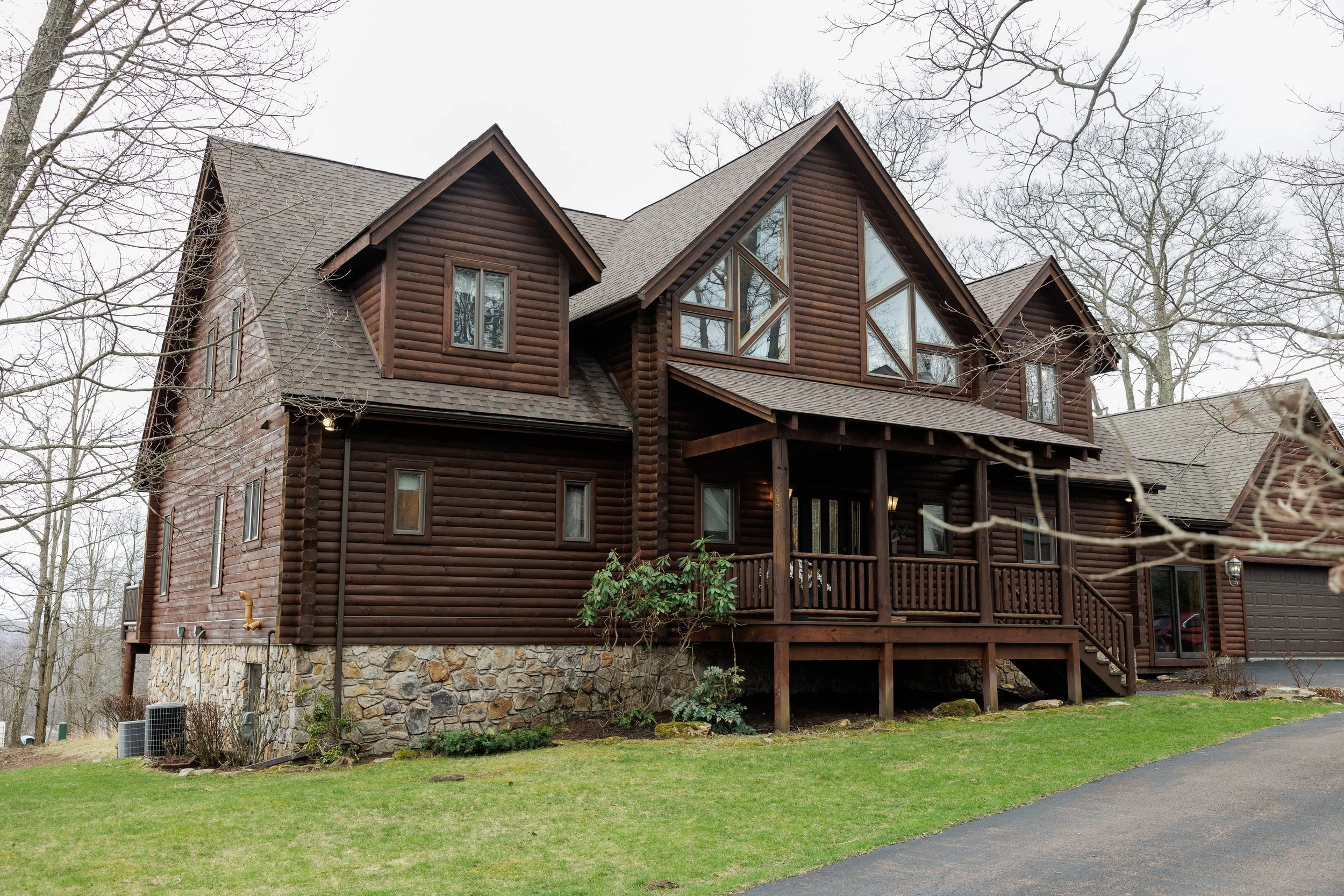 A large wooden house with a stone foundation, multiple gabled roofs, and large triangular windows, set in a wooded area with leafless trees and a grassy yard.