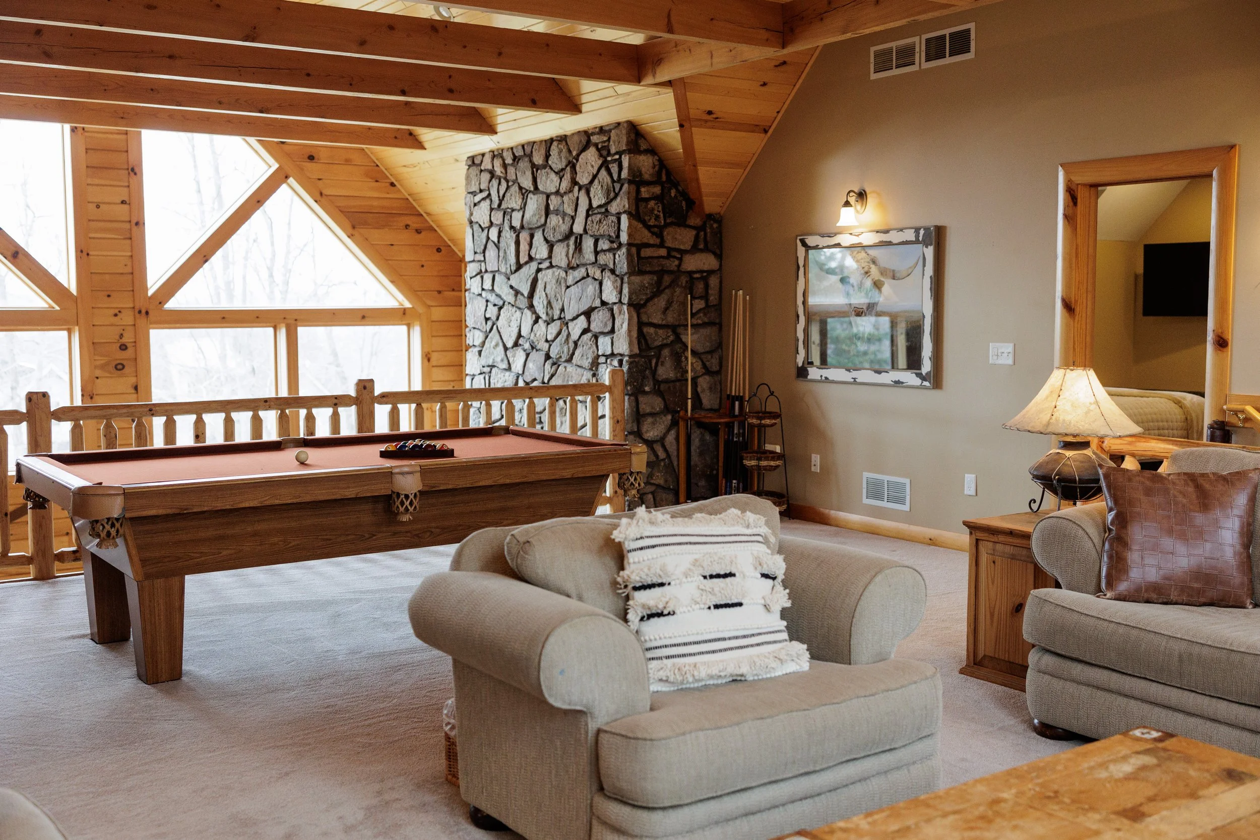 Living room with wooden ceiling and walls, featuring a stone fireplace, a pool table, a mirror on the wall, a lamp, and comfortable chairs with throw blankets.