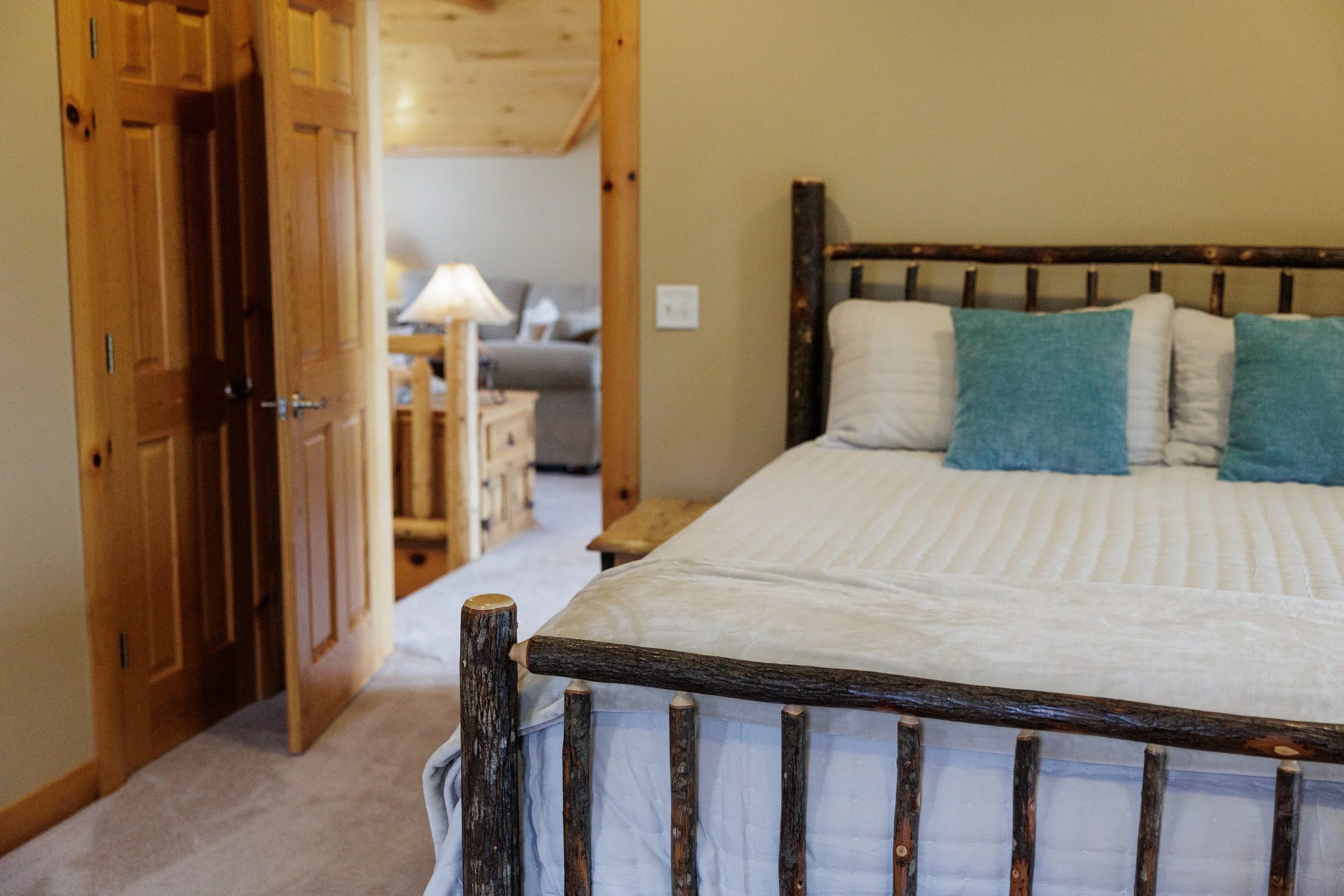 A bedroom with a rustic log bed, beige bedding, and blue pillows. In the background, a doorway leads to a cozy living area with a lamp on a wooden table and a sofa.