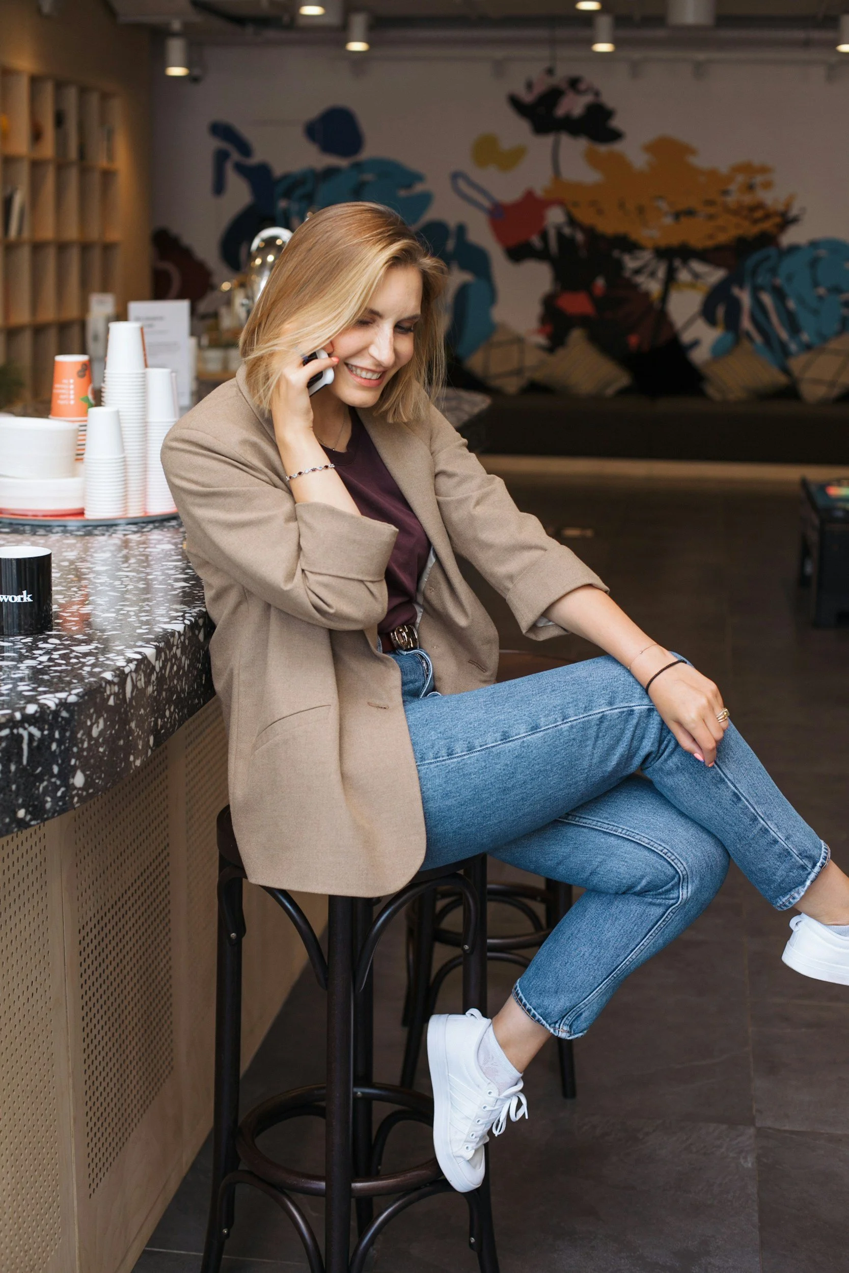 A young woman with blonde hair, wearing a beige blazer, maroon top, and blue jeans, sitting on a bar stool in a cafe, smiling and talking on her mobile phone. In the background, there is colorful abstract wall art.
