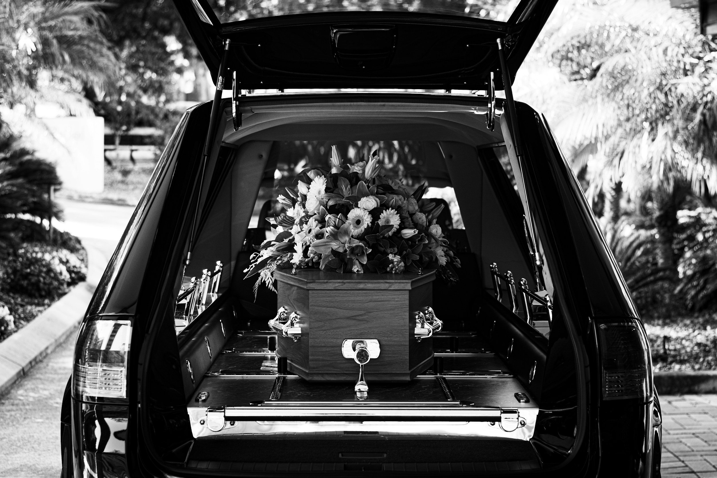 Black hearse with an open rear door filled with a floral casket, parked outdoors on a paved area.