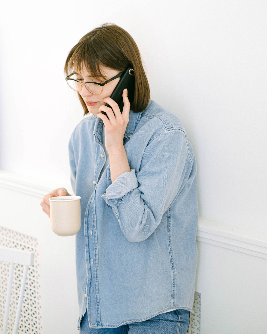 A woman with shoulder-length brown hair, glasses, and a denim shirt is talking on the phone and holding a white mug in a bright room.