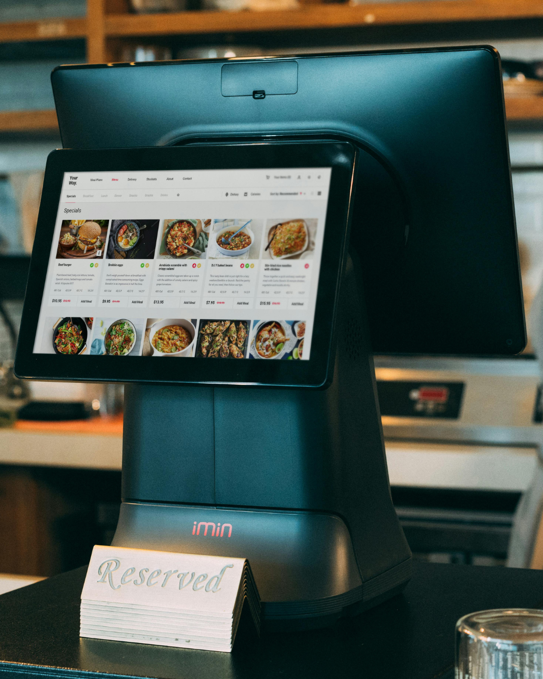 Touchscreen ordering kiosk with a digital menu displaying various food dishes, positioned on a black counter with a reserved sign in front.