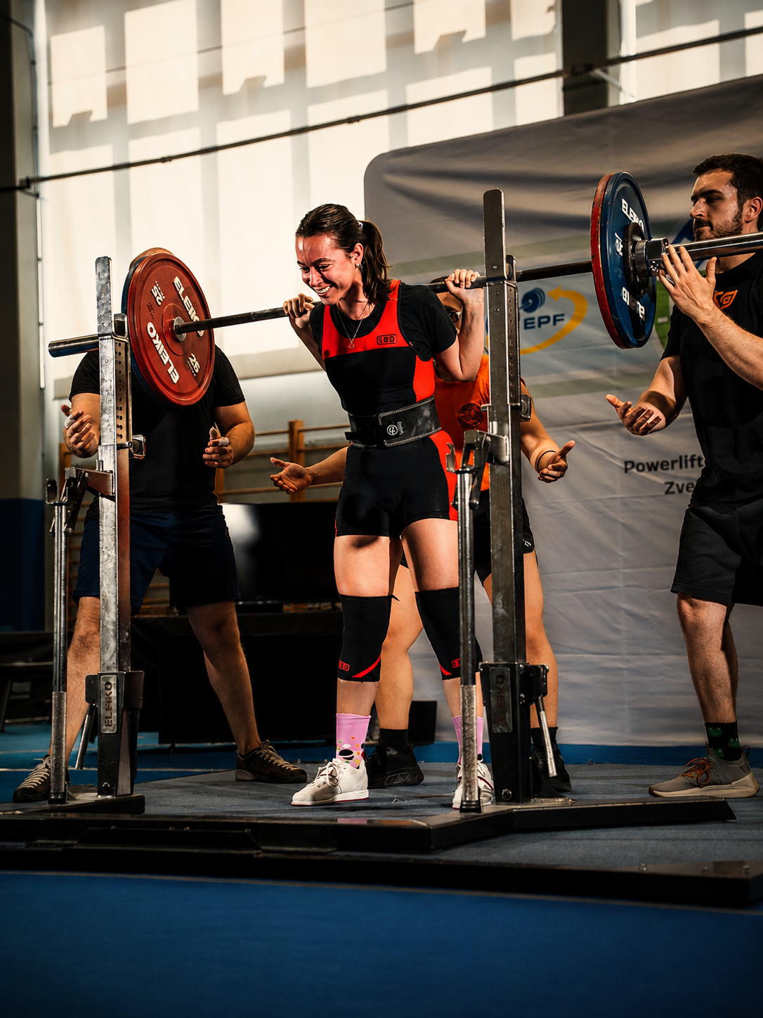 Donna che solleva un bilanciere durante una competizione di powerlifting, circondata da altri partecipanti o allenatori in un ambiente di palestra.