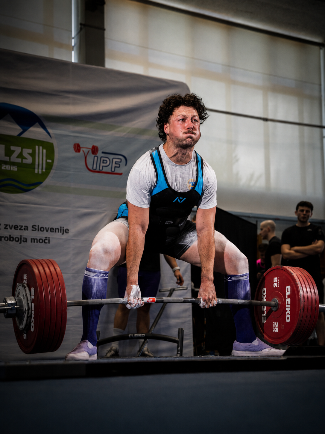Uomo che solleva un bilanciere con pesi rossi durante una competizione di sollevamento pesi indoor, con diversi spettatori sullo sfondo e uno sfondo con logo e scritte in sloveno.