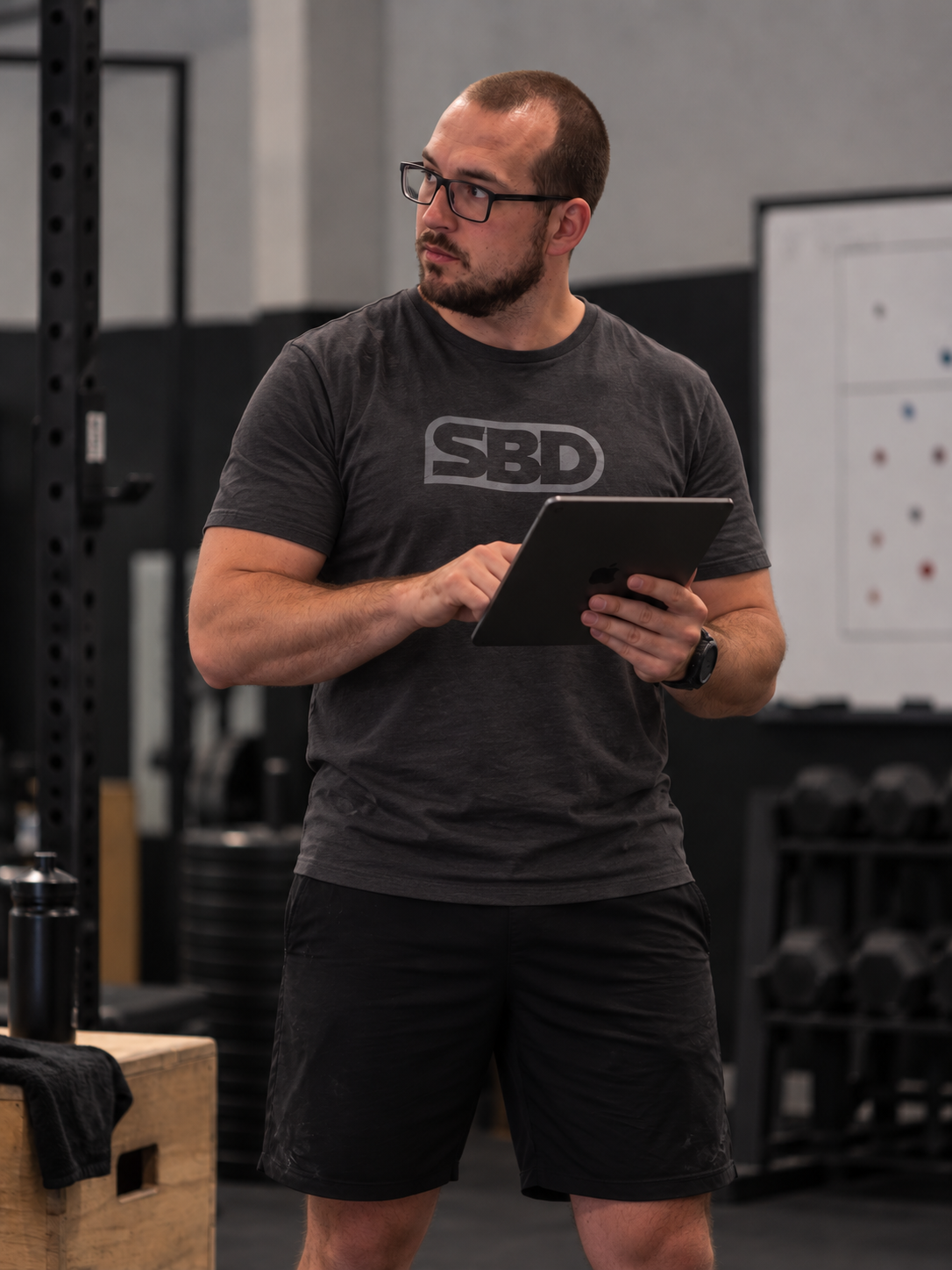 Un uomo con occhiali, barba corta e vestito sportivo nero che tiene un tablet in una palestra, con attrezzi da fitness e una parete con una lavagna bianca sullo sfondo.