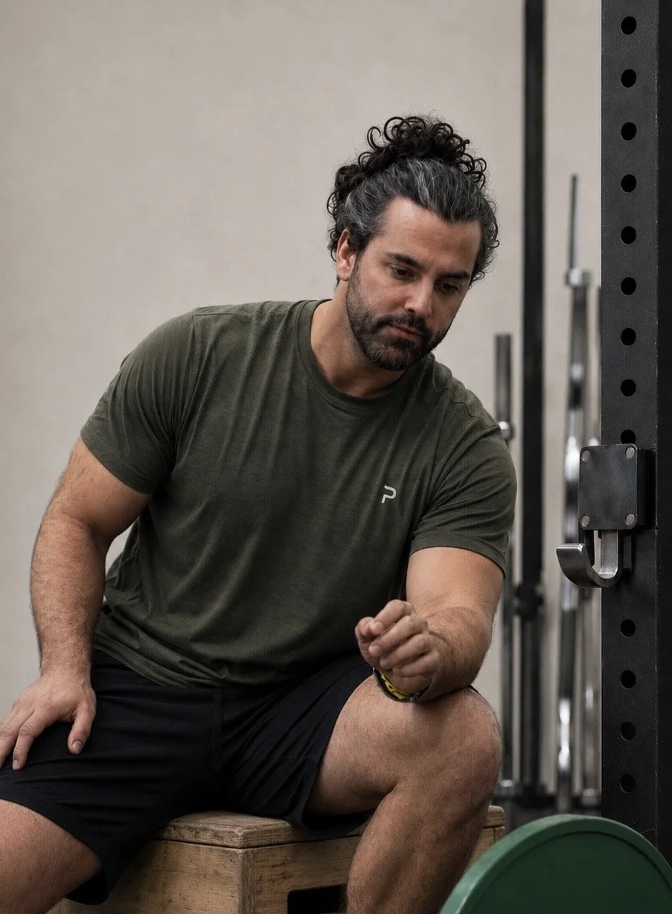Uomo che fa stretching o riscaldamento prima dell'allenamento in palestra, seduto su una panca di legno con attrezzature di allenamento sullo sfondo.