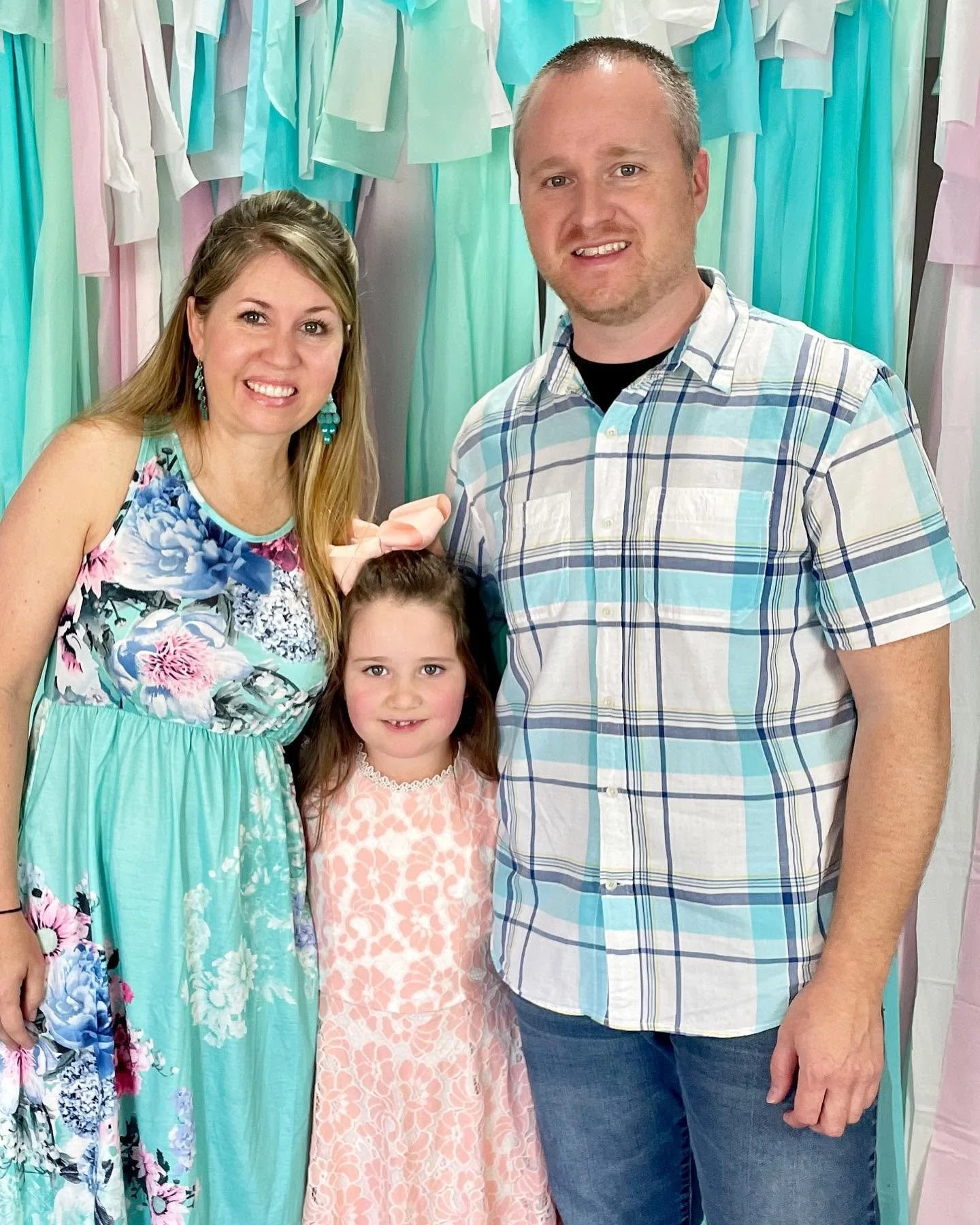 A family of three standing in front of a colorful backdrop made of pastel-colored fabric strips. The woman on the left is wearing a floral dress, the girl in the middle is wearing a pink dress with a large pink bow in her hair, and the man on the right is wearing a blue plaid shirt and jeans.