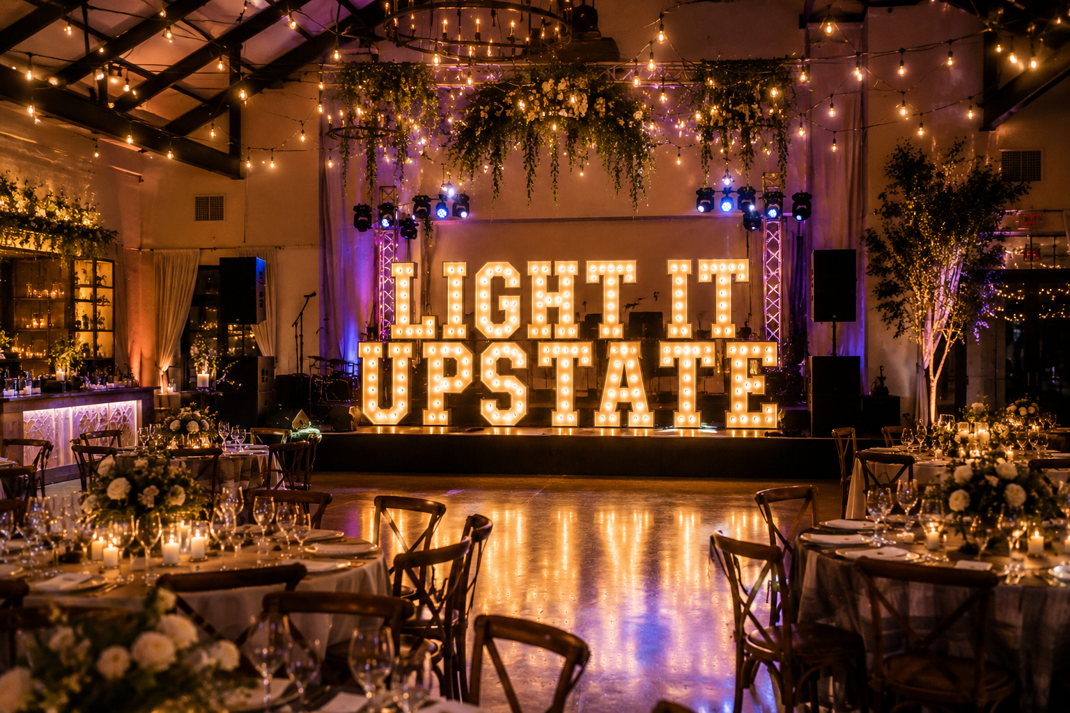 A decorated event space with a stage featuring large illuminated marquee letters spelling 'Light It Up State,' surrounded by string lights, floral arrangements, and tables set for a celebration.