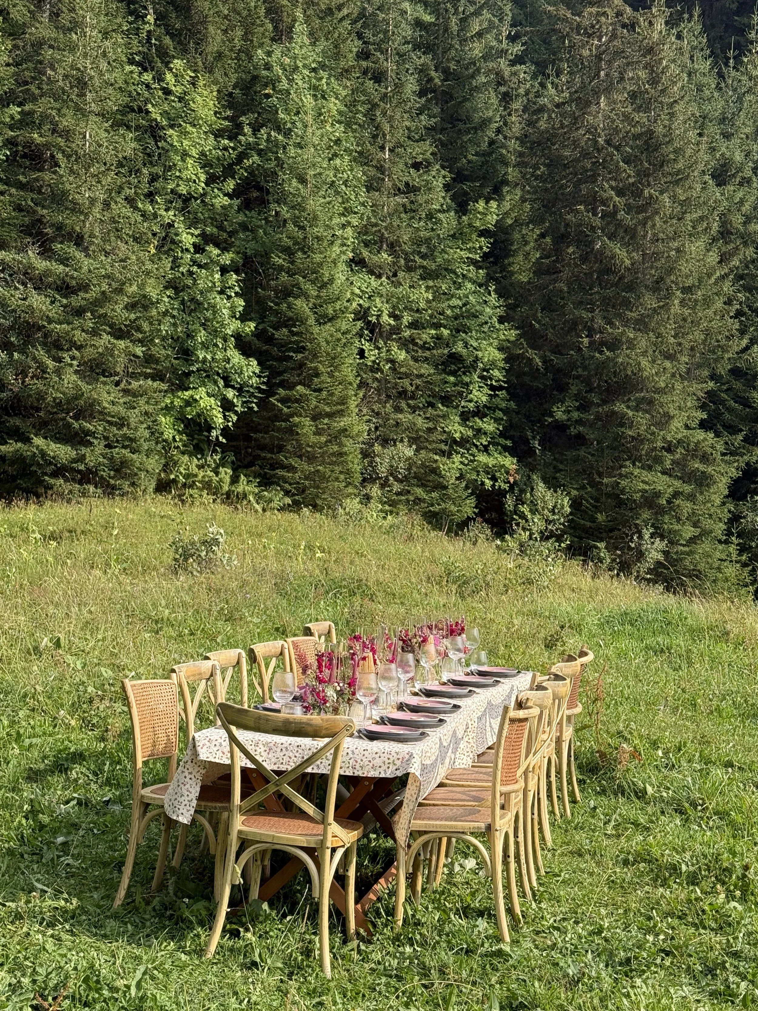 An outdoor dining table set in a grassy clearing surrounded by trees, with chairs, plates, glasses, and floral centerpieces.
