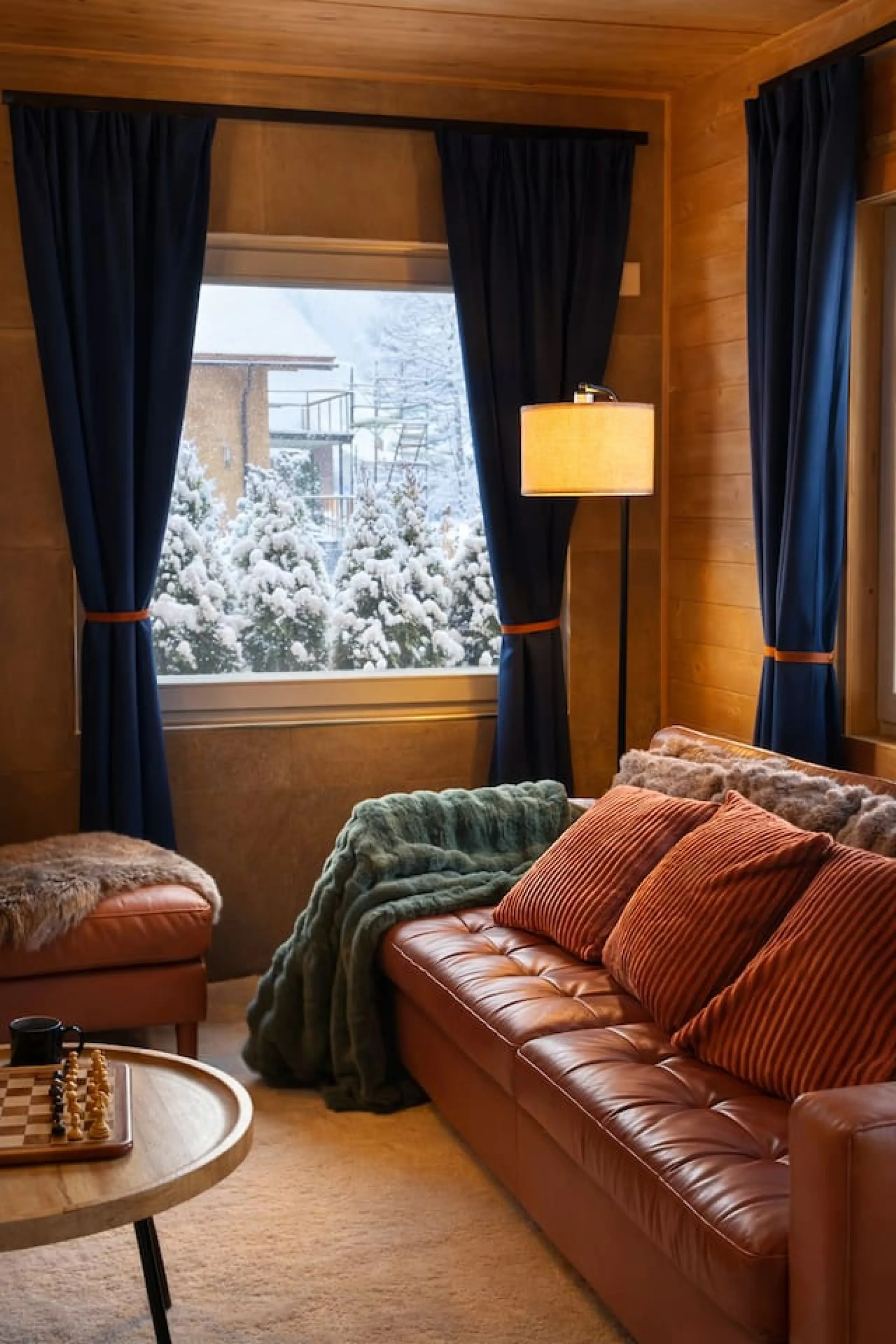 View of a cozy living room with a large window overlooking a snow-covered landscape, blue curtains, a floor lamp, a leather couch with striped pillows, a matching ottoman with a fur throw, and a round wooden table with a chess set.