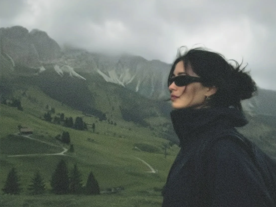 A woman with black hair and sunglasses looking to the side, standing outdoors with a mountain landscape and cloudy sky in the background.
