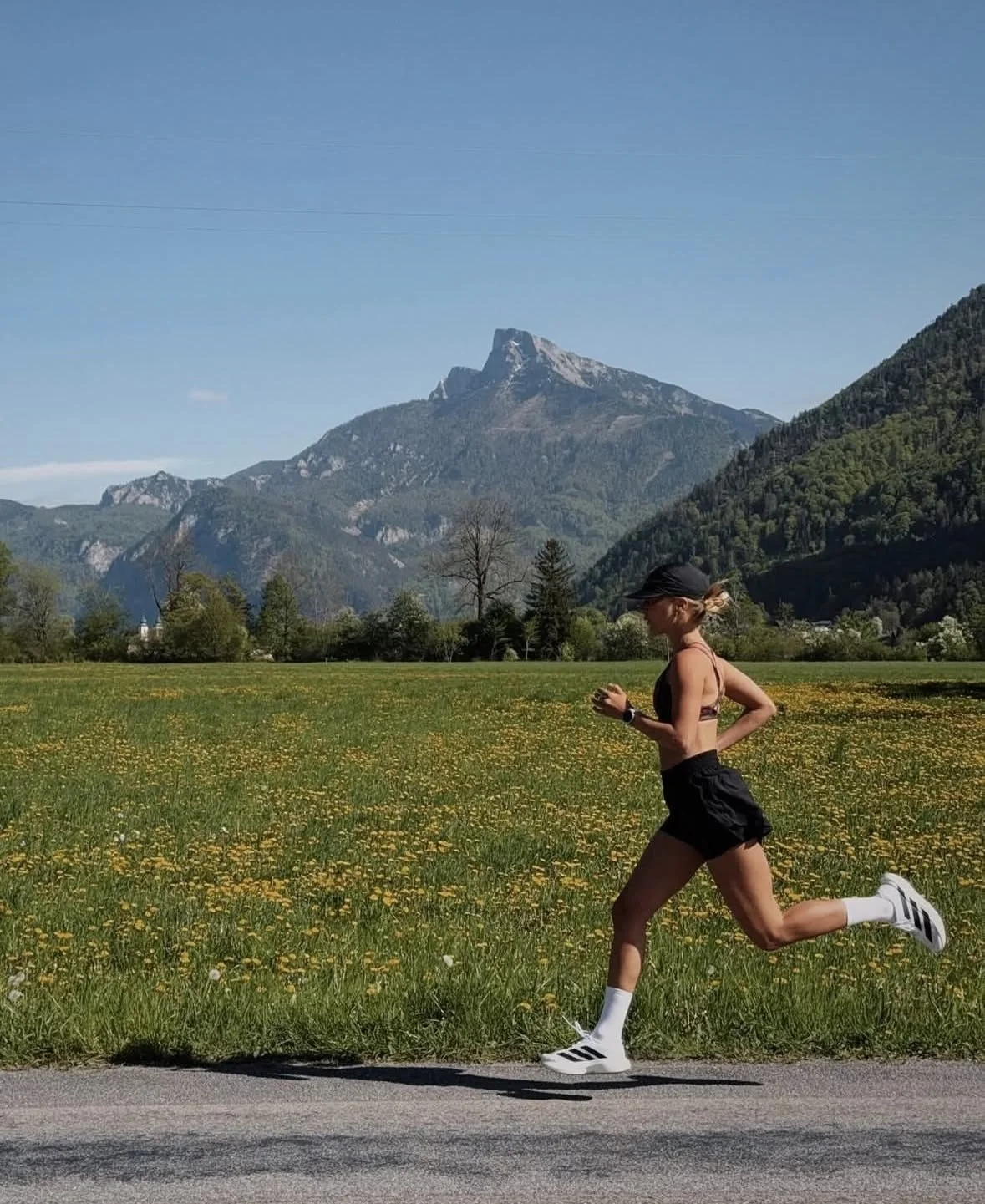 A woman jogging on a paved road in a grassy field with yellow flowers, mountains in the background under a clear blue sky.