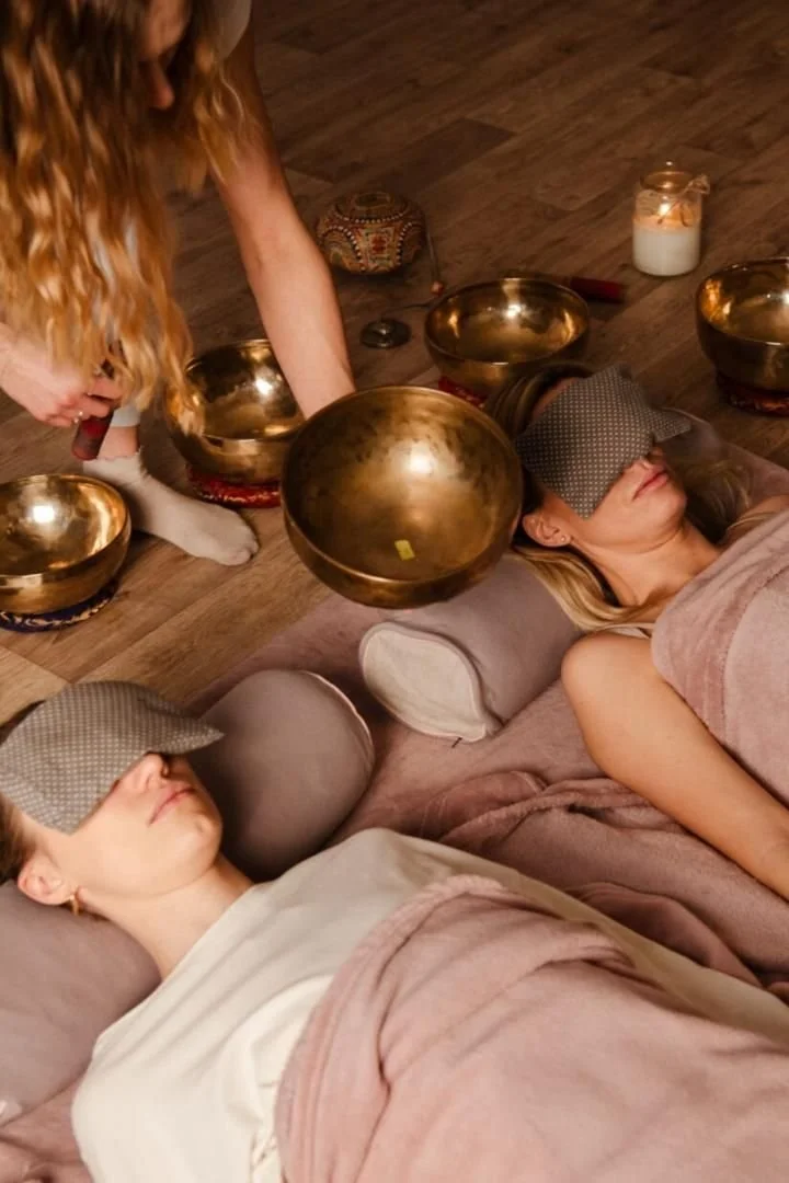 Two women lying down with meditation pillows, blindfolded, receiving sound therapy from a person playing singing bowls, in a cozy setting.