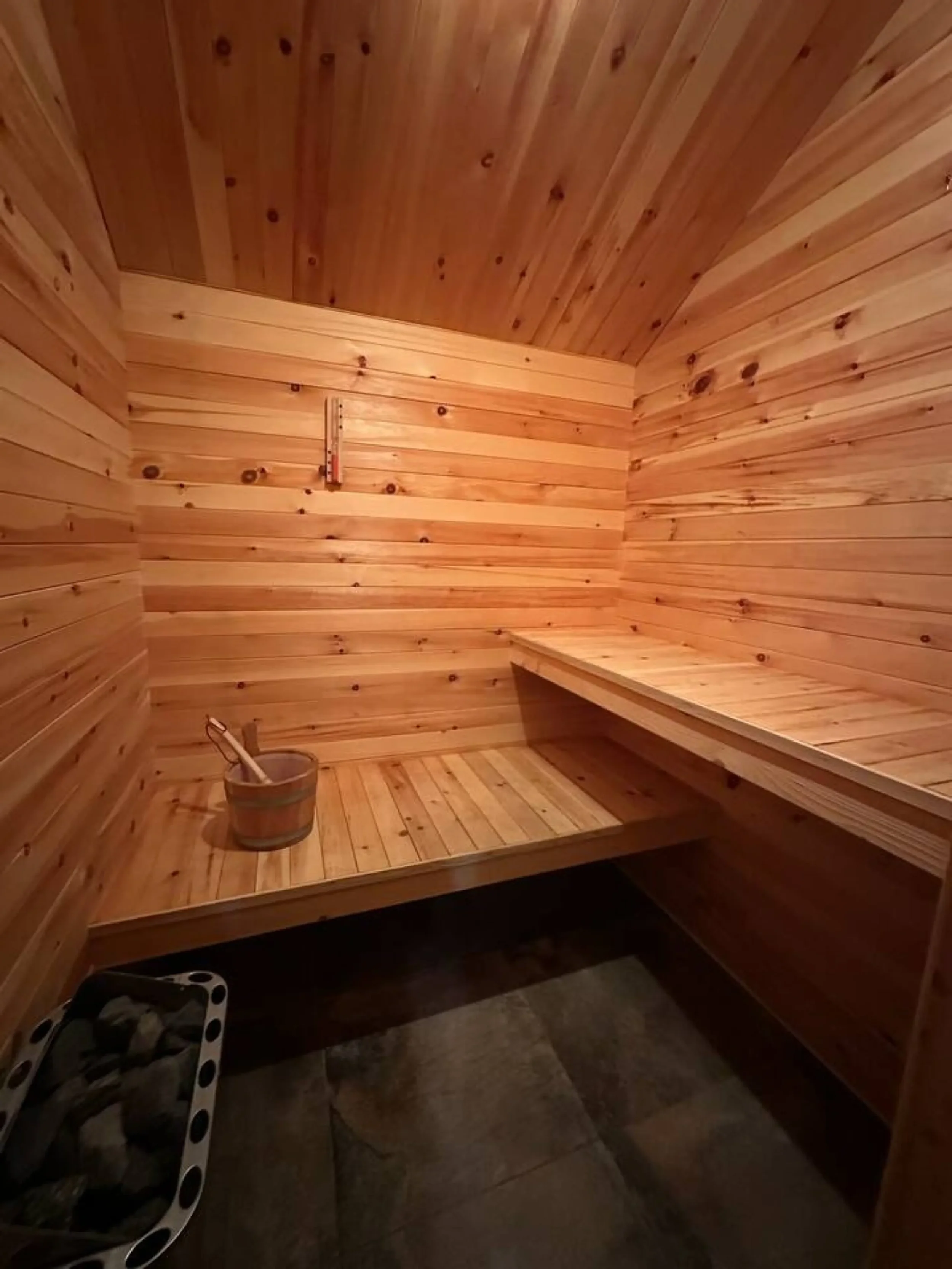 A small wooden sauna room with wood-paneled walls and ceiling, a bench along the back wall, a door on the left side, and a bucket with a ladle on the floor, with a stone heater in the corner.