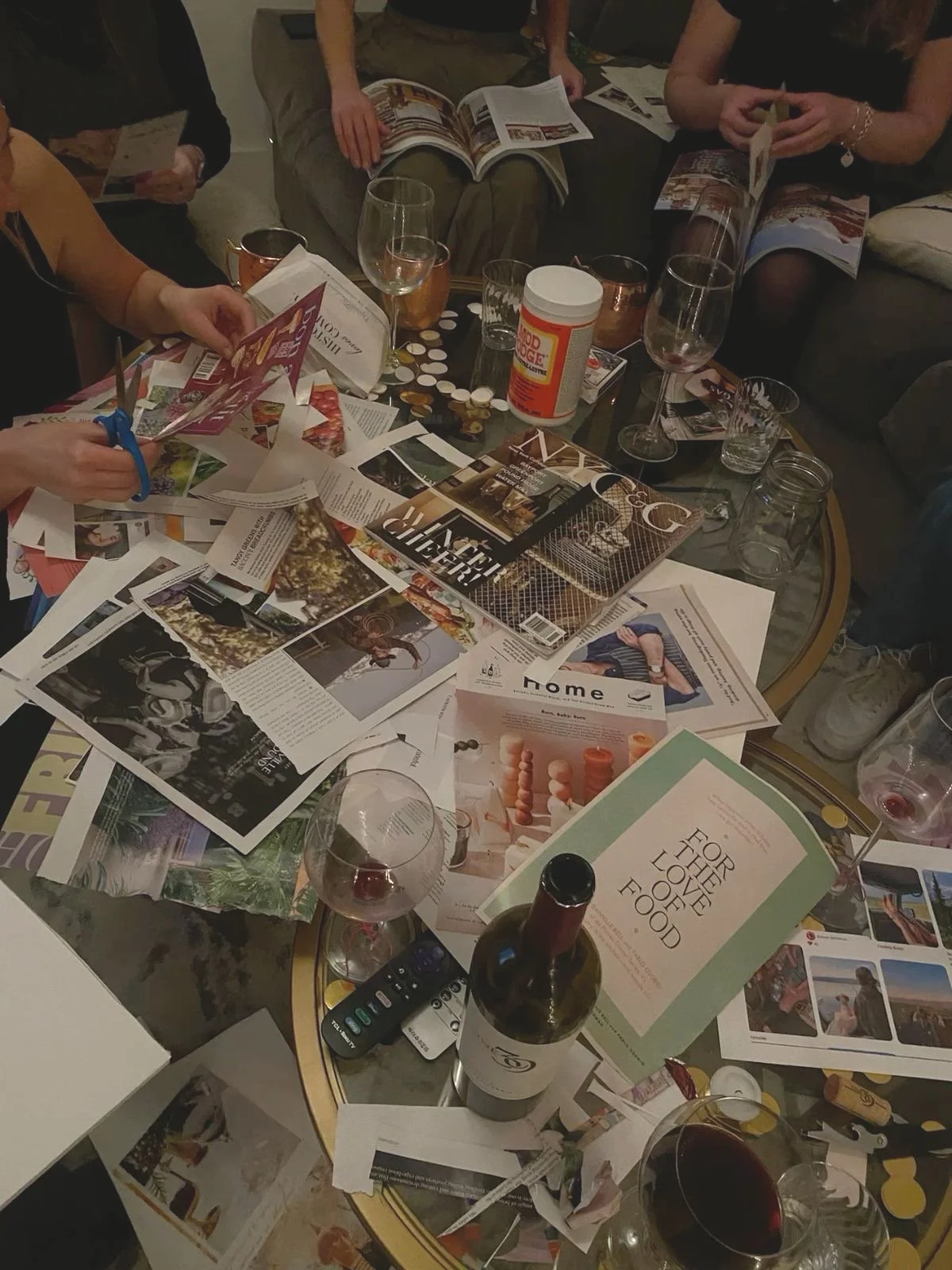 A cluttered glass coffee table covered with magazines, brochures, scattered papers, wine glasses, a bottle of wine, a remote control, and other miscellaneous items, with people sitting around it.