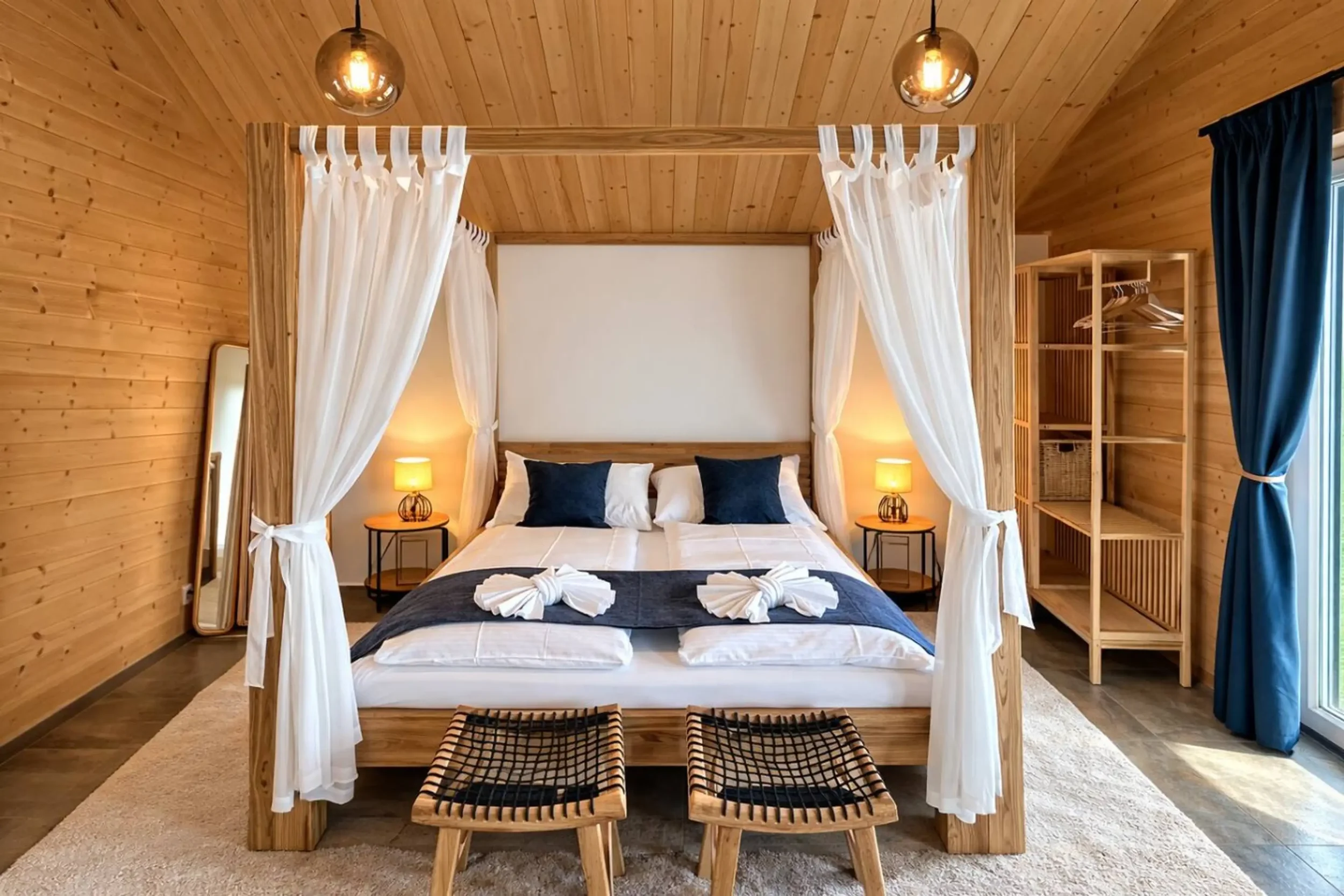 A bedroom with a wooden canopy bed, white canopy curtains, and blue and white bedding, illuminated by two bedside lamps and ceiling lights, with a wooden wardrobe and mirror on the side.