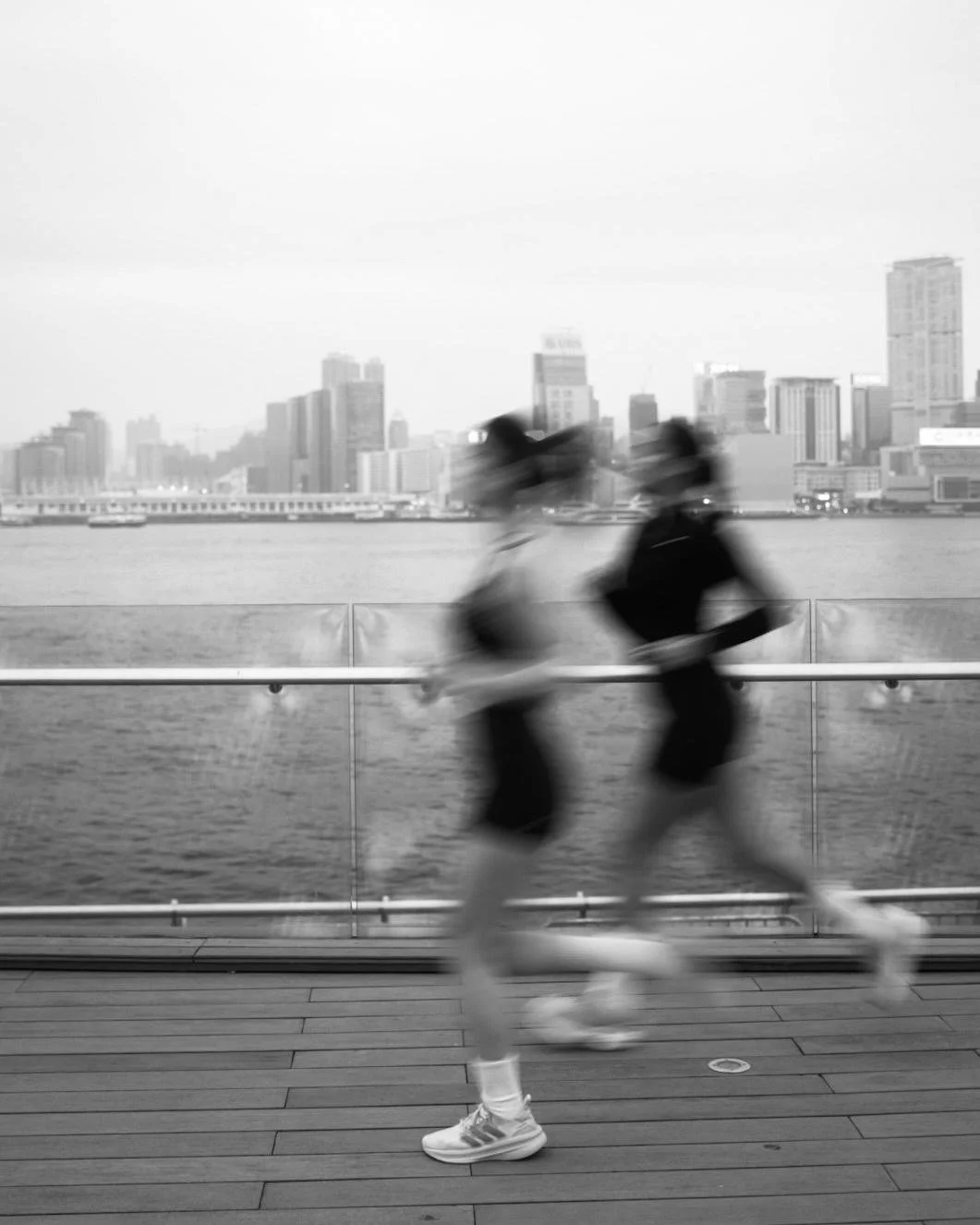 Black and white photo of a runner in motion on a waterfront promenade with a city skyline in the background.
