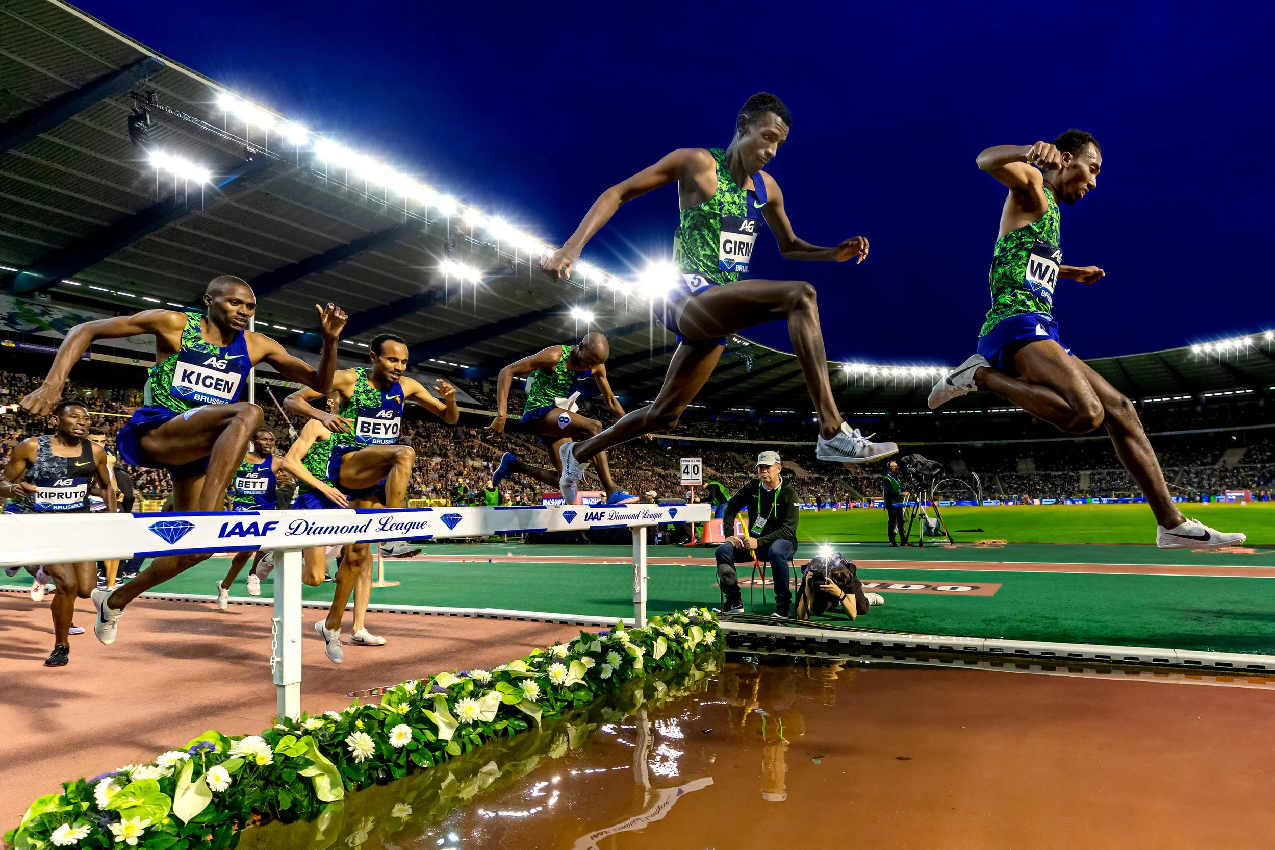 Track and field athletes start a race on an indoor stadium track with a water hazard in front, spectators in the stands, and bright stadium lights overhead.