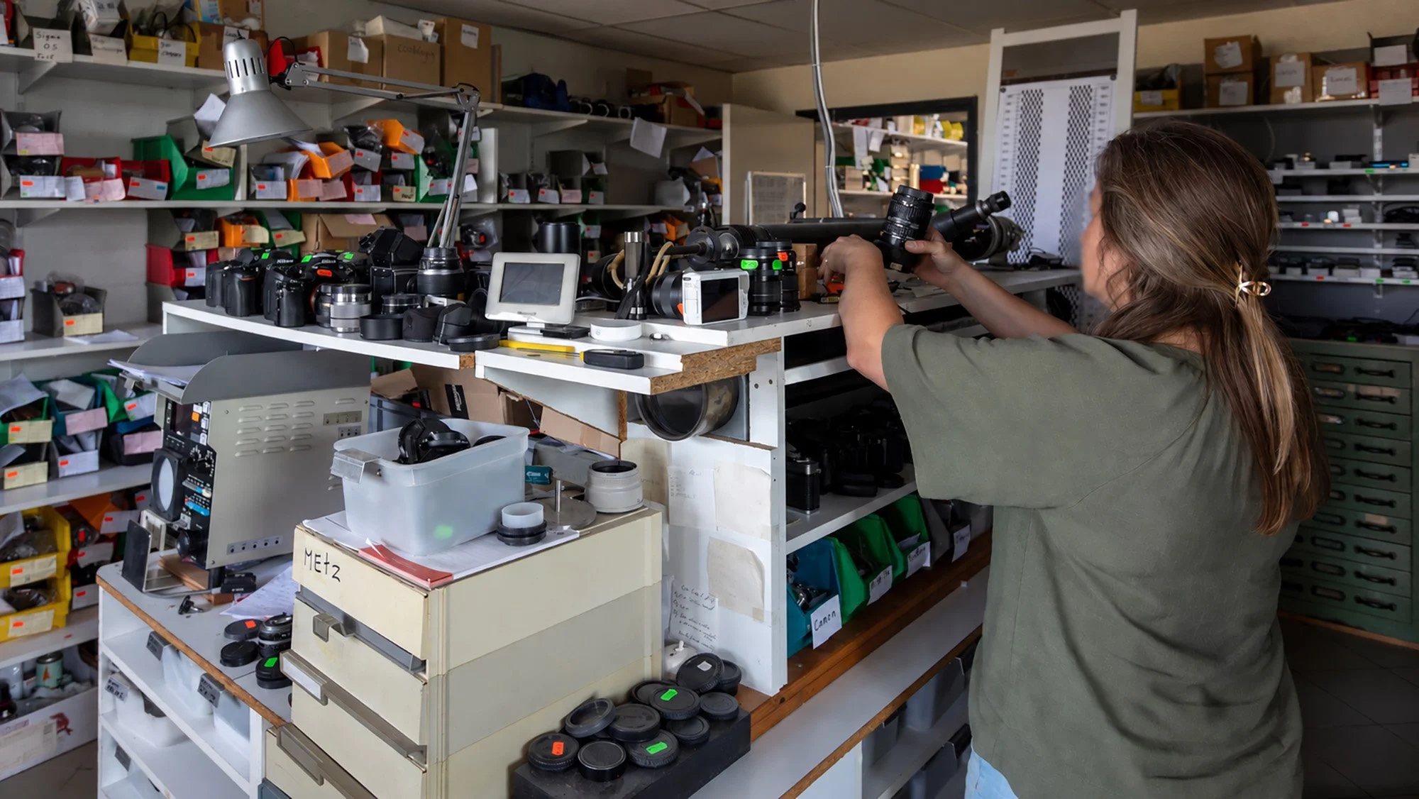 A woman working at a cluttered table filled with camera equipment, lenses, and accessories in a storage room with shelves of labeled boxes.