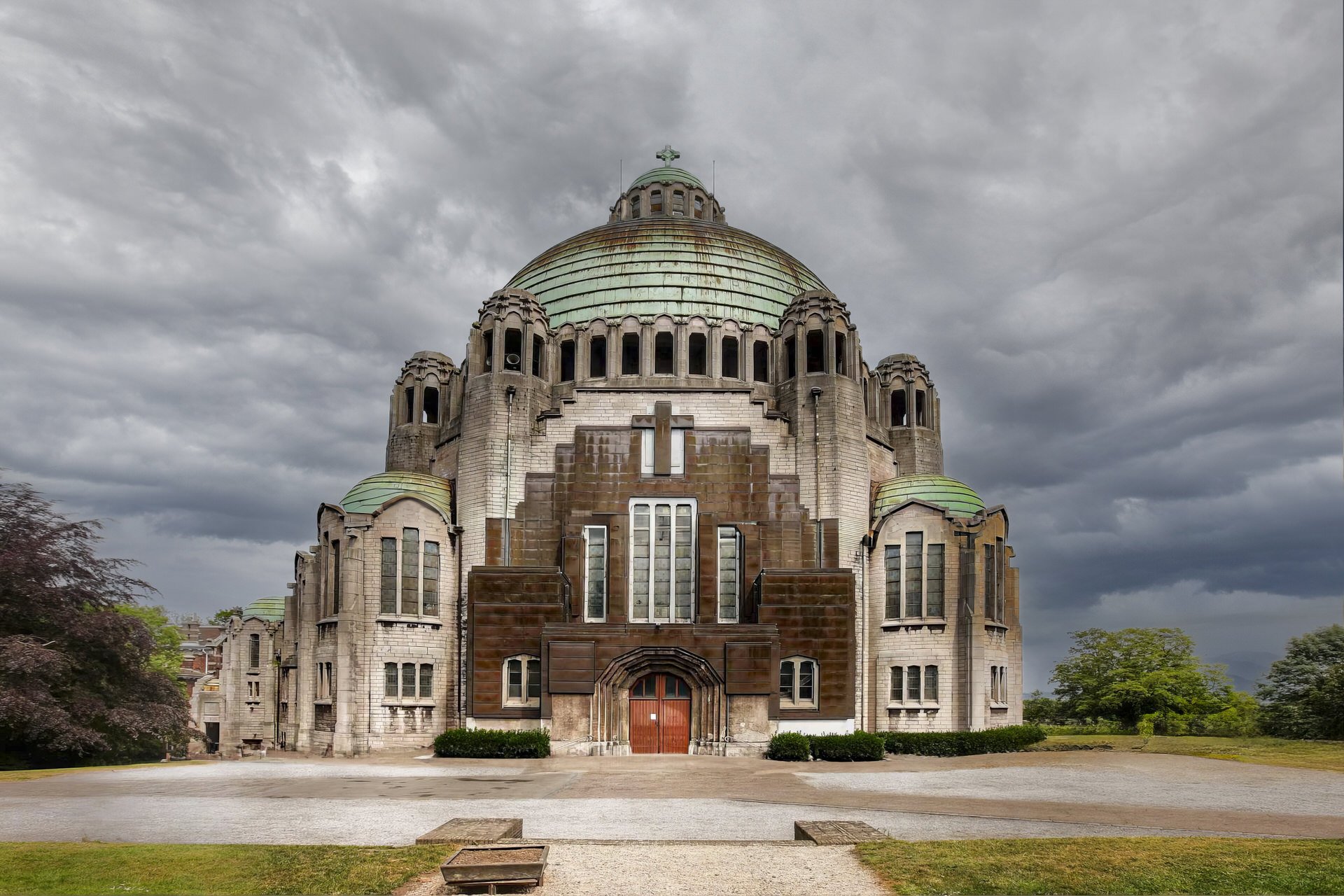 A large historic church with a prominent green copper dome, stone walls, and arched windows, set against a cloudy sky with green trees and a gravel pathway in front.