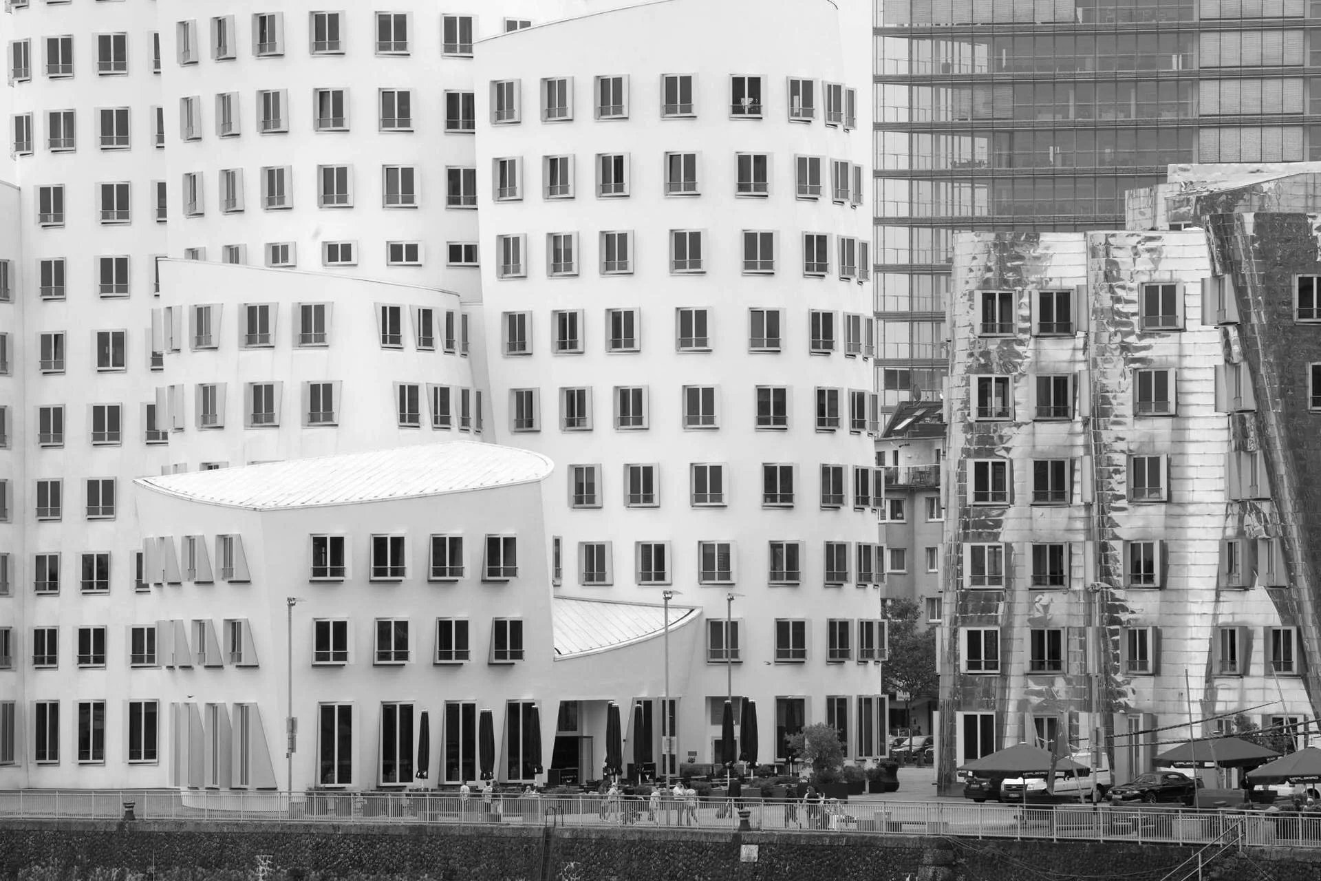 Black and white photo of modern glass and concrete buildings with many windows and small balconies, along a street with parked cars and pedestrians, in an urban cityscape.