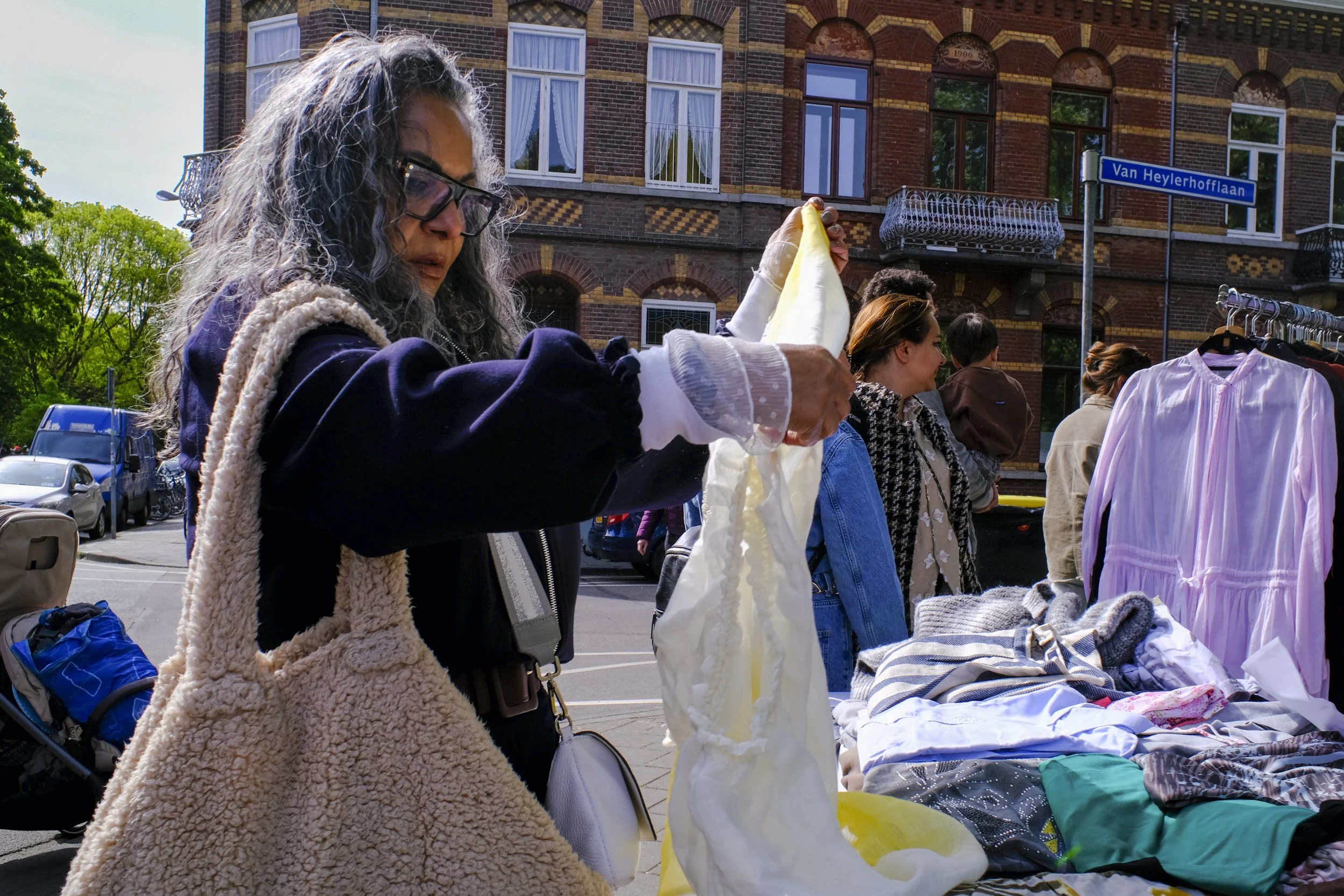 A woman with curly gray hair and glasses browsing clothes at an outdoor market stall on Van Heylerhofflaan in front of a brick building with arched windows. Other people are browsing nearby, and tables are filled with various clothes and textiles.