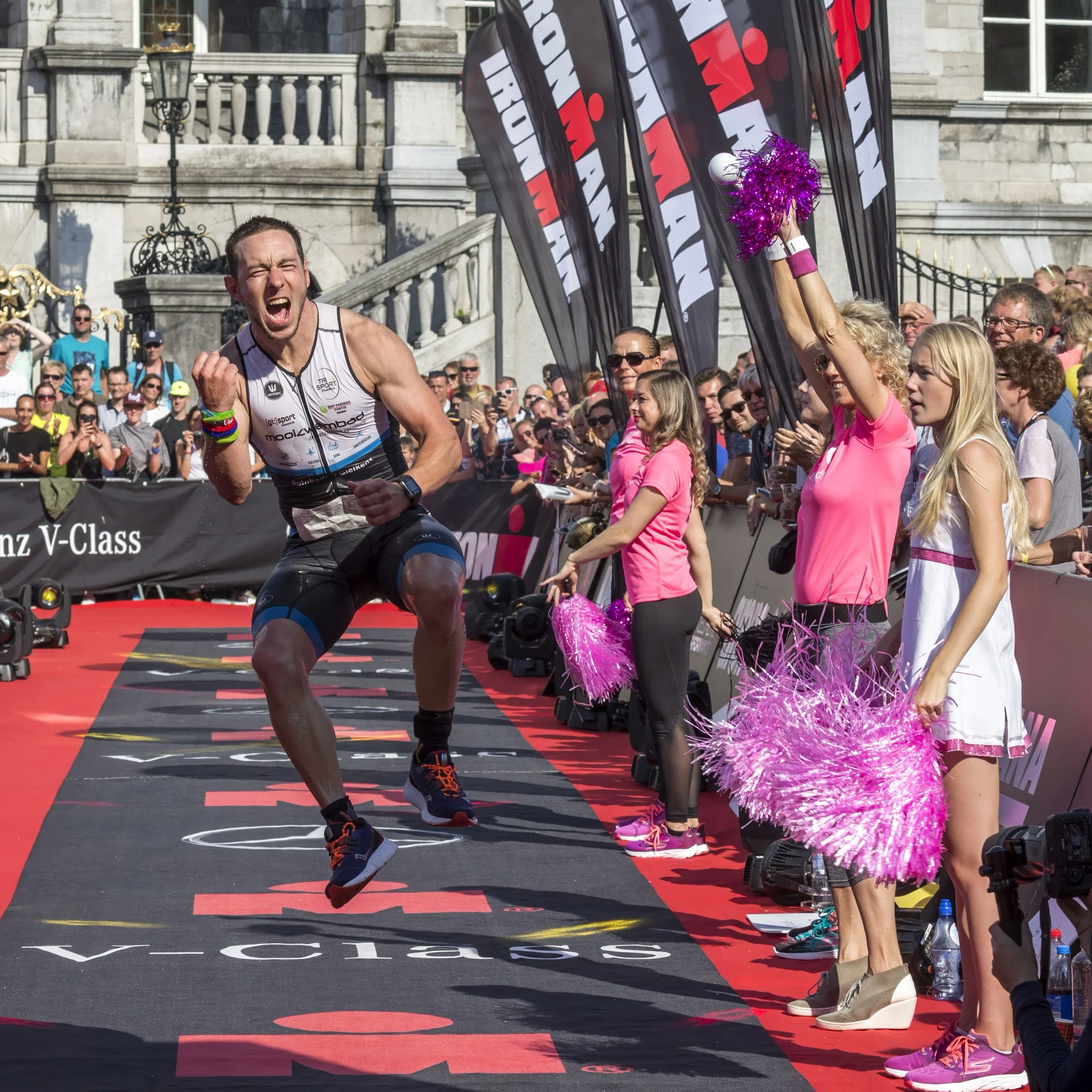 A male athlete crossing the finish line at a race event, surrounded by spectators and supporters, with bright pink cheerleading pom-poms held by a woman in pink shirt and glasses.