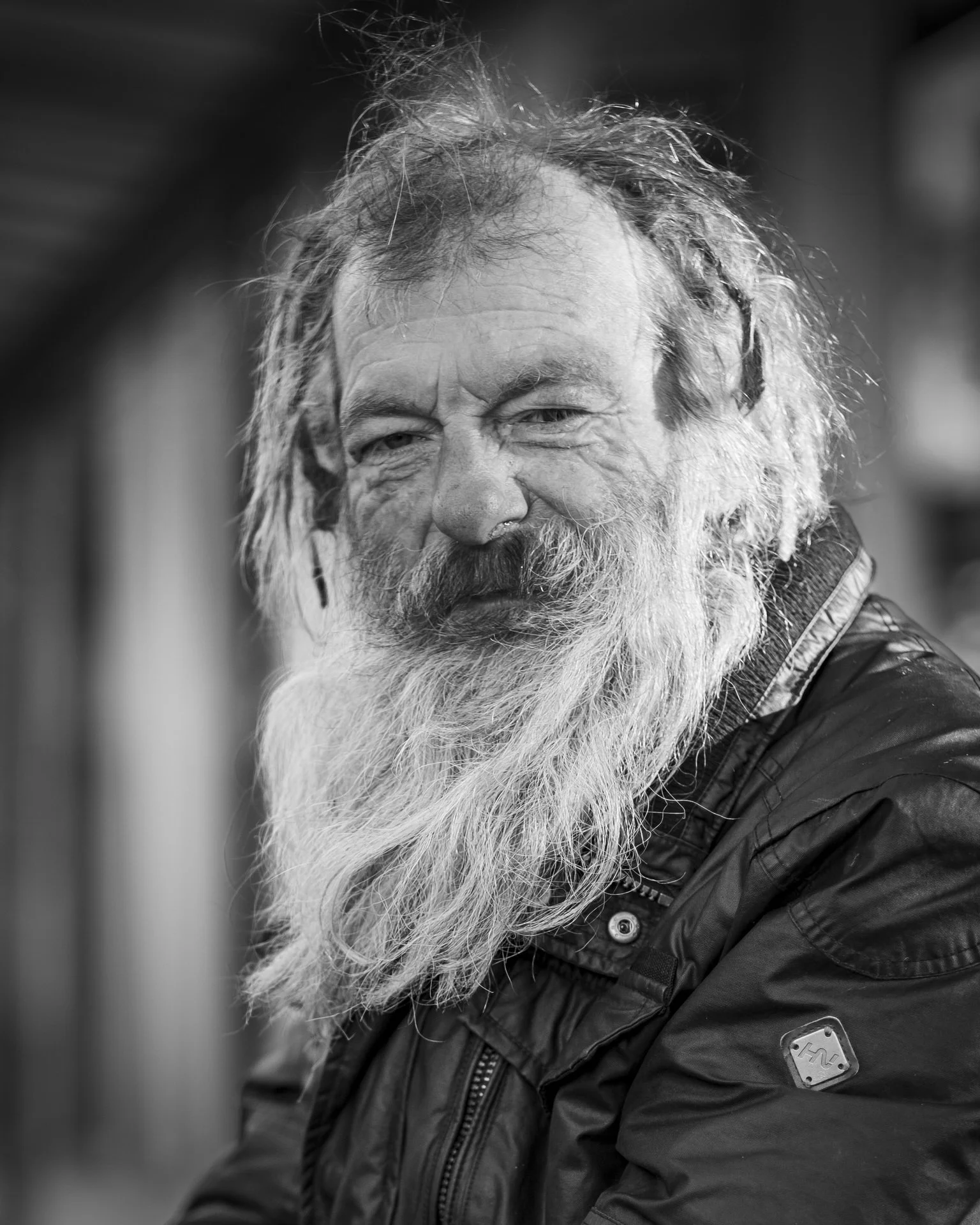 Close-up black and white photograph of an older man with long, unkempt hair and a full beard, wearing a dark jacket with a patch on the sleeve.