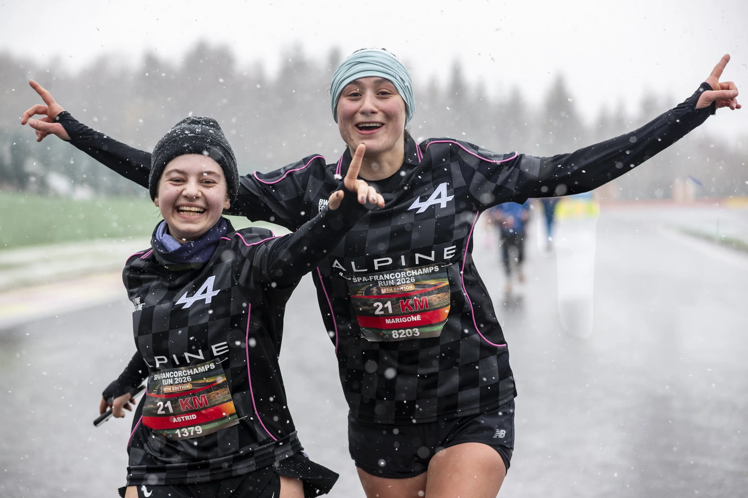 Two women running outdoors in rainy weather, smiling and celebrating, wearing black running attire with race bibs, part of a charity or marathon event.