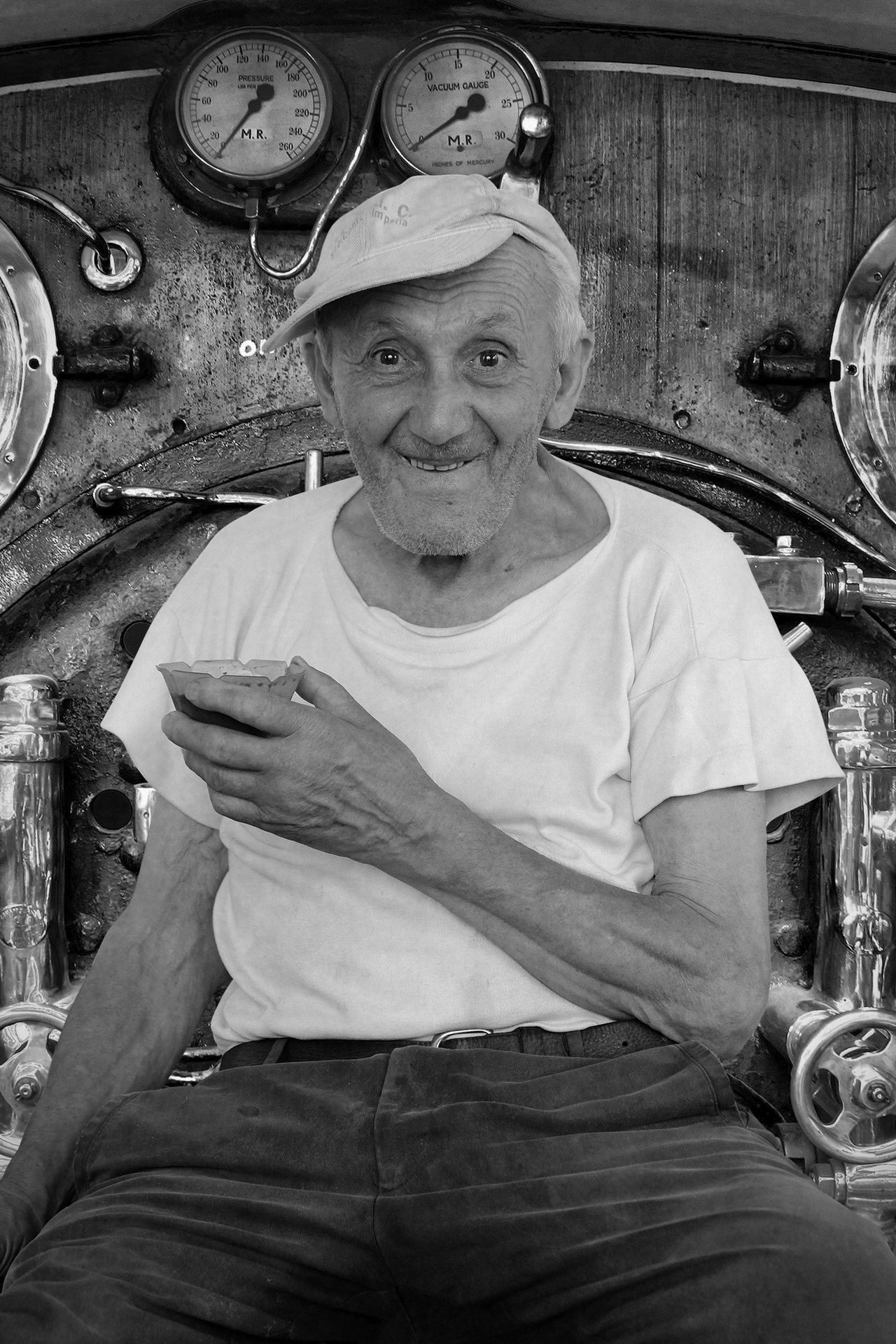 An elderly man with a beard, wearing a cap backwards and a white t-shirt, sitting in front of a vintage pressure and vacuum gauge panel, smiling and holding a small bowl in his right hand.