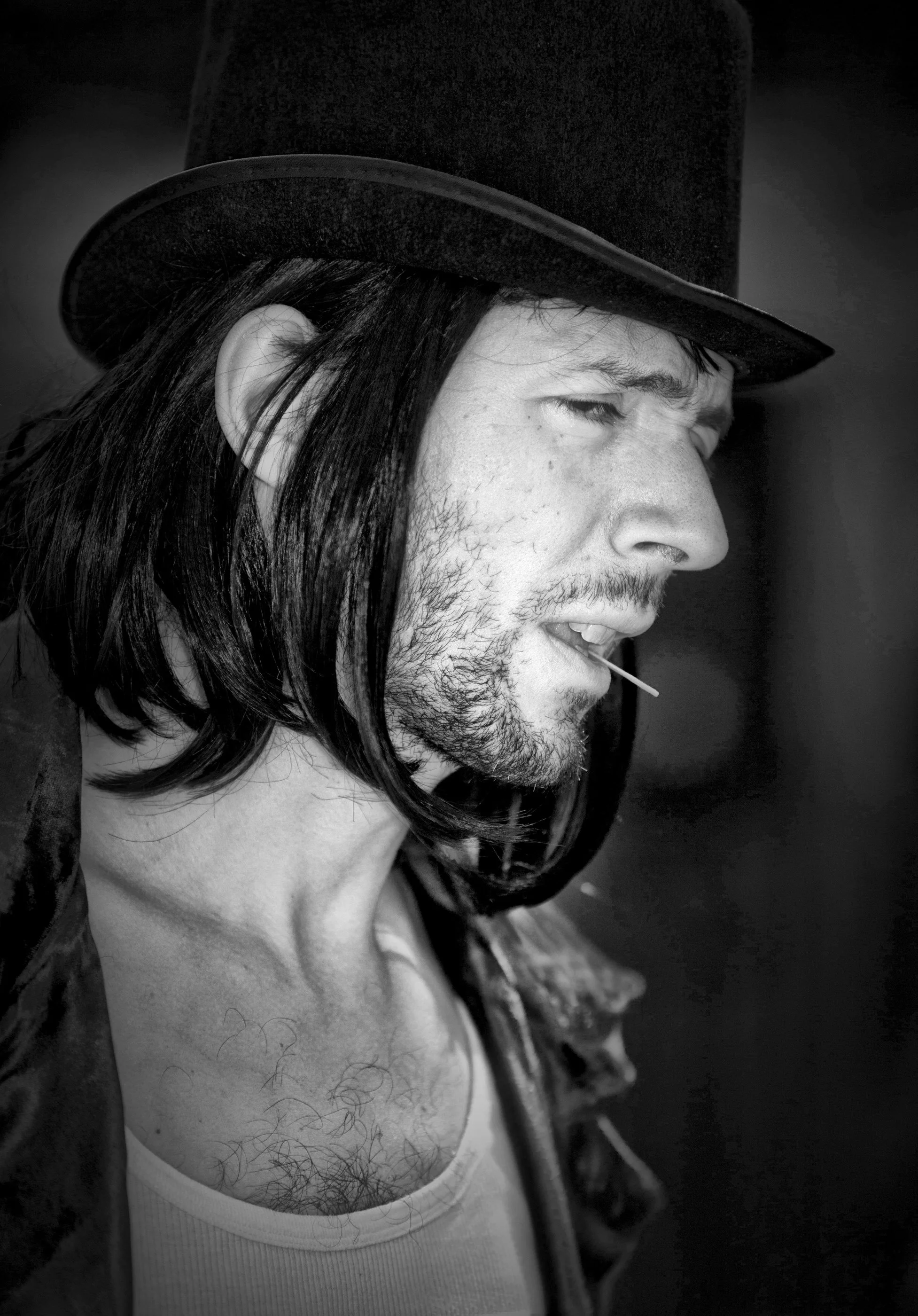 Black and white close-up of a man with long hair, a beard, and a mustache wearing a top hat and a white tank top with visible chest hair, holding a toothpick in his mouth.