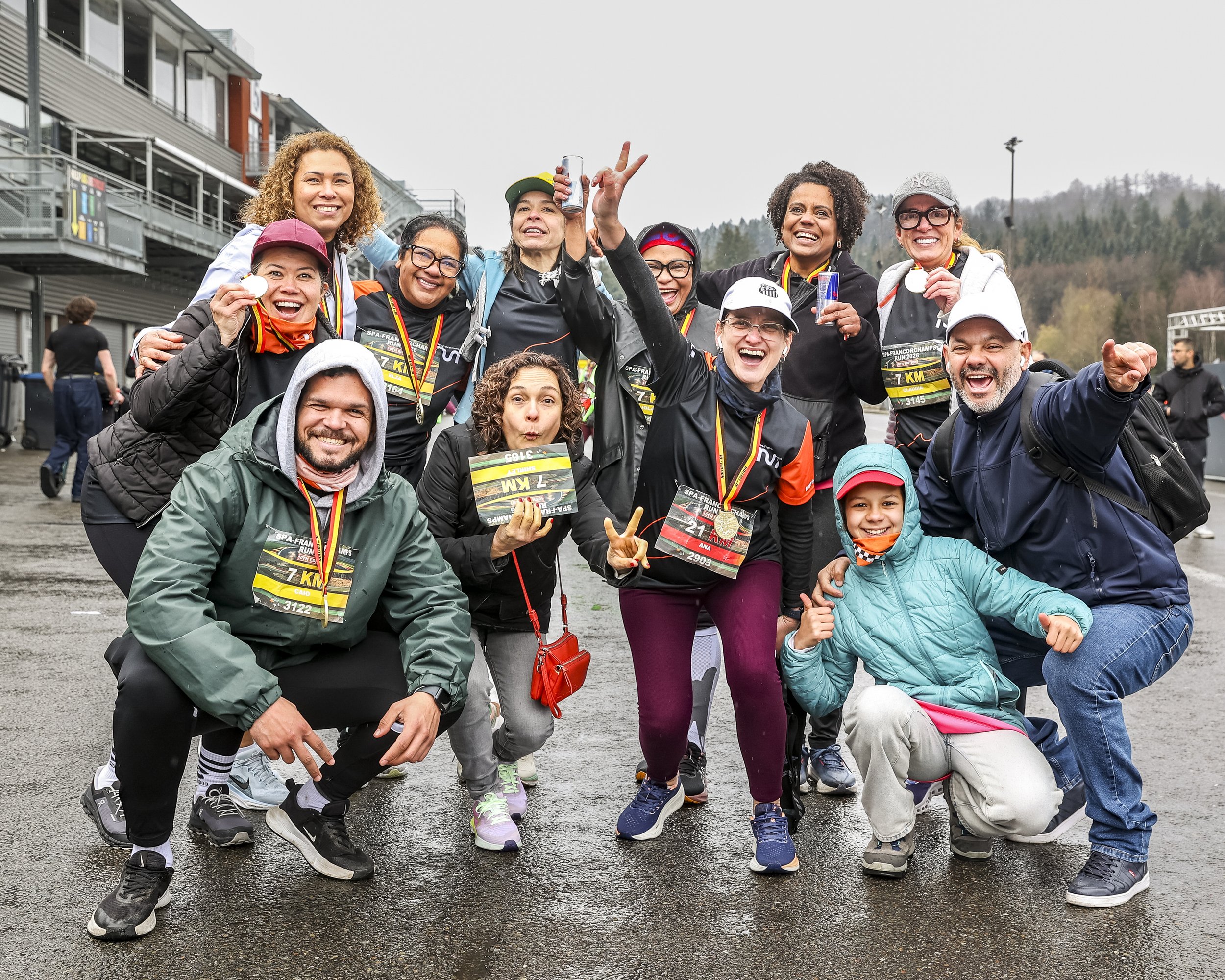 A group of runners celebrating after finishing a race outdoors on a cloudy day, wearing bibs and medals, with some making victory signs and smiling.