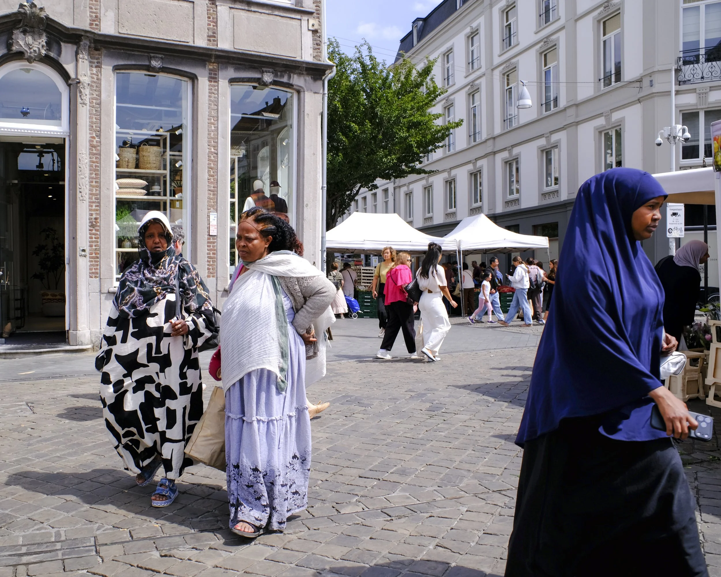 People walking on cobblestone street in an urban area with white buildings, tents, and trees.