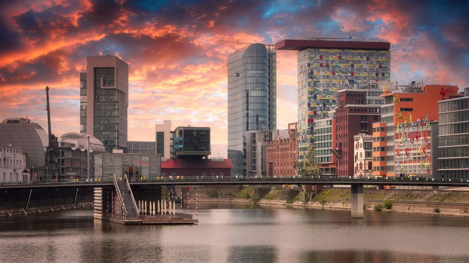 City skyline during sunset with modern skyscrapers, colorful sky, and a river in the foreground.
