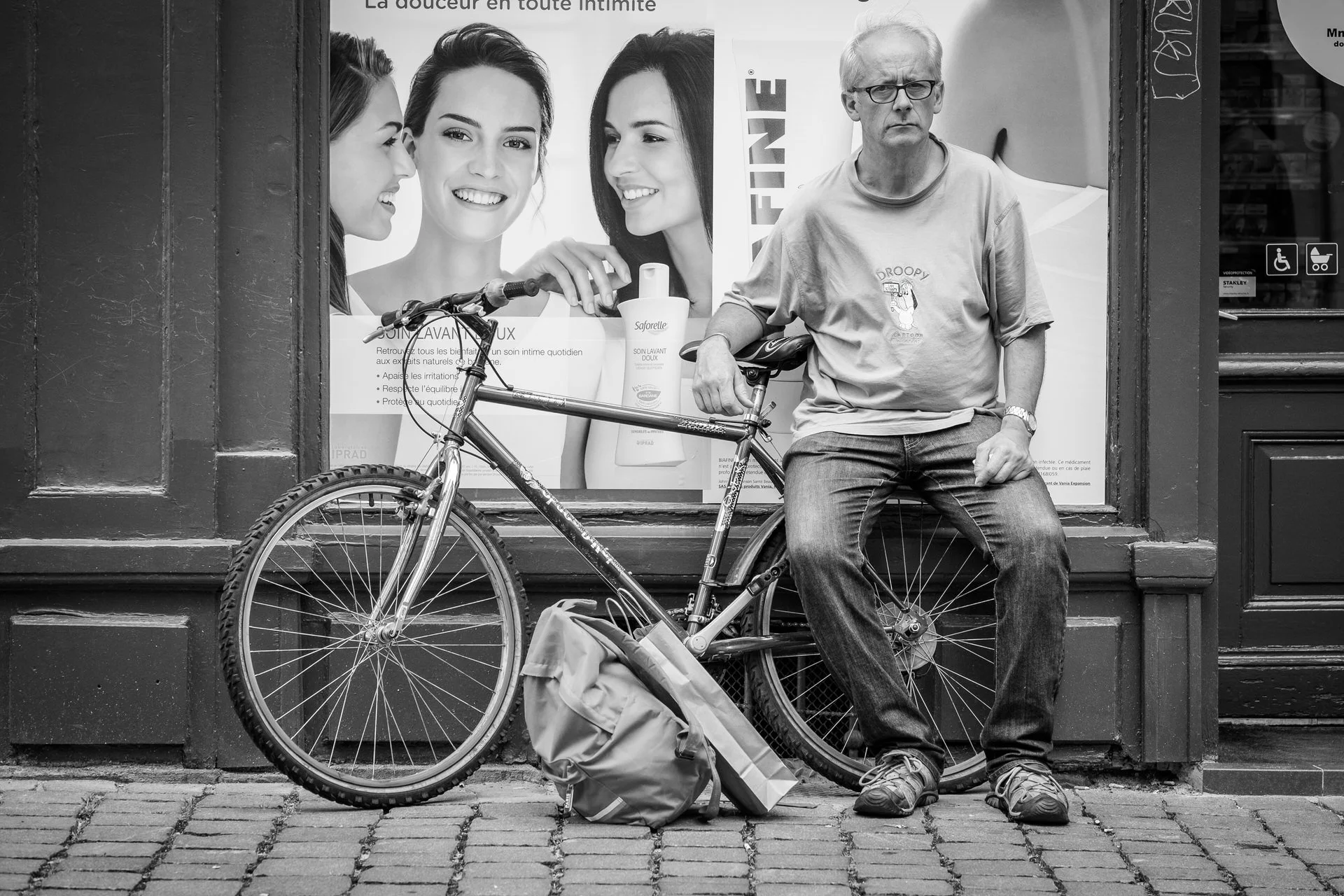 A man with glasses and a t-shirt sits on a bicycle in front of a storefront window. The window has a large poster showing three smiling women and skincare products. The bicycle leans against the storefront, and there is a bag on the ground in front o