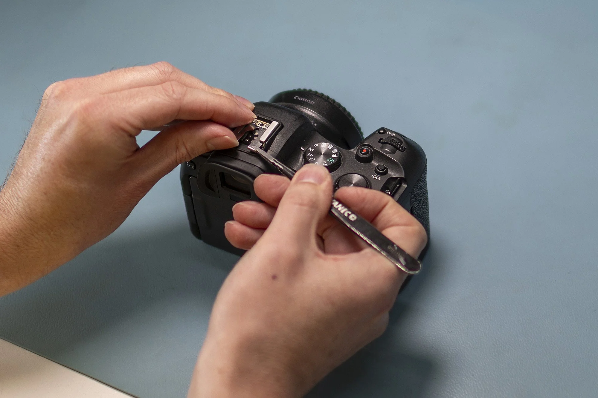 Hands holding a DSLR camera and a screwdriver, assembling or repairing the camera on a blue surface.